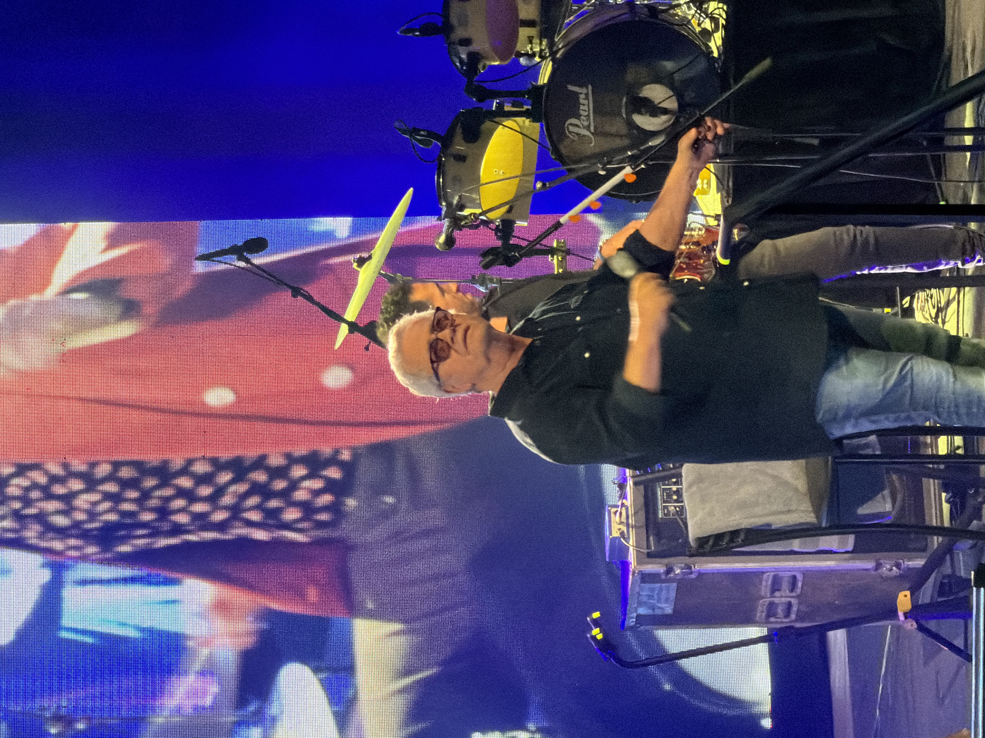 The image depicts a man, presumably a drummer, performing on stage in Tel Aviv, Israel. He is positioned behind a drum kit, identifiable by the "Pearl" logo on the bass drum. The stage lighting creates a blue hue in the background. A large screen behind the drummer displays a close-up of a man's face. Stage equipment is visible in the foreground.