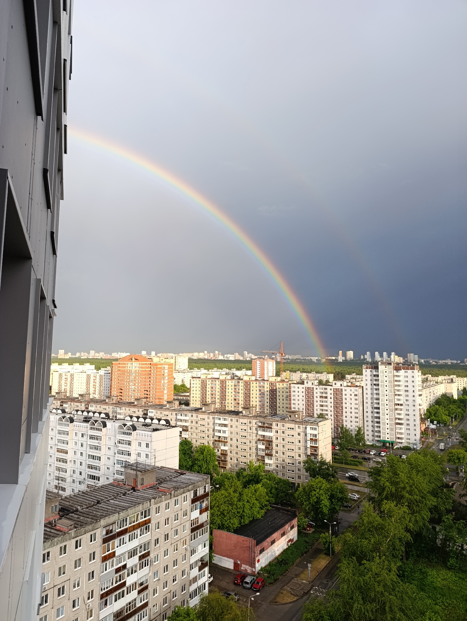 The image shows a cityscape of Perm, Russia under a dramatic sky featuring a double rainbow. The foreground is dominated by densely packed, mostly white and gray apartment buildings with flat roofs. Some buildings show signs of age with discolored facades. Green trees are interspersed among the buildings, especially towards the lower right corner of the frame. Cars are visible on a street in the lower right. In the background, more apartment buildings are visible, along with a construction crane and some industrial structures. The sky is gray, transitioning from light gray where the rainbow originates to a darker, stormier gray towards the right side. The double rainbow arches over the cityscape, its colors clearly visible. A portion of the building from which the photo was taken is visible on the left side. The scene suggests a post-rain environment with a mix of urban development and natural elements.