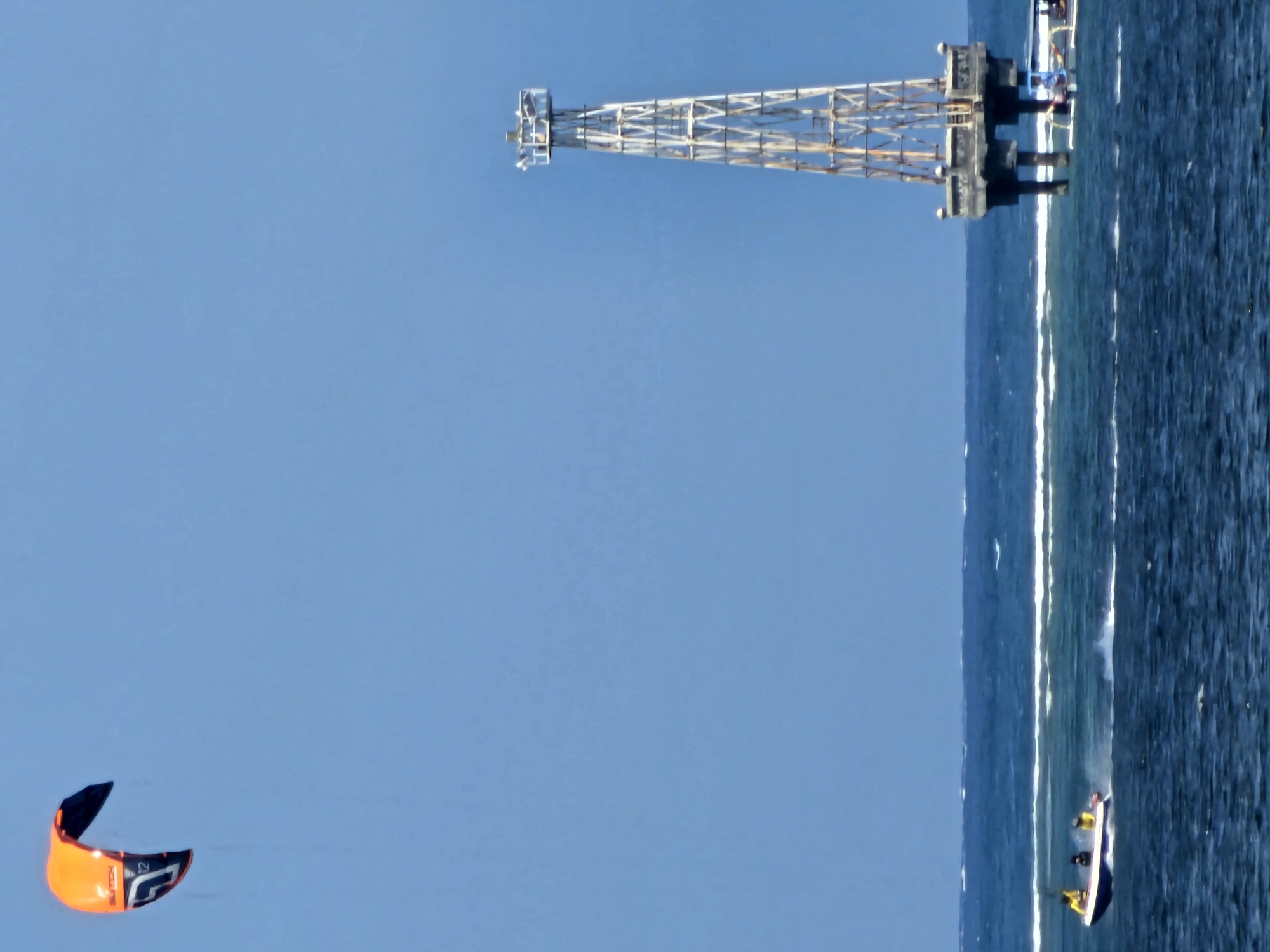 An orange and black kitesurfing kite flies in a clear blue sky. In the background, a metal tower structure with a platform at its base stands in the ocean. A small boat with three people on it is moving across the water. The scene appears to be a coastal area, possibly near Denpasar, Indonesia, given the water conditions and the presence of recreational water activities.