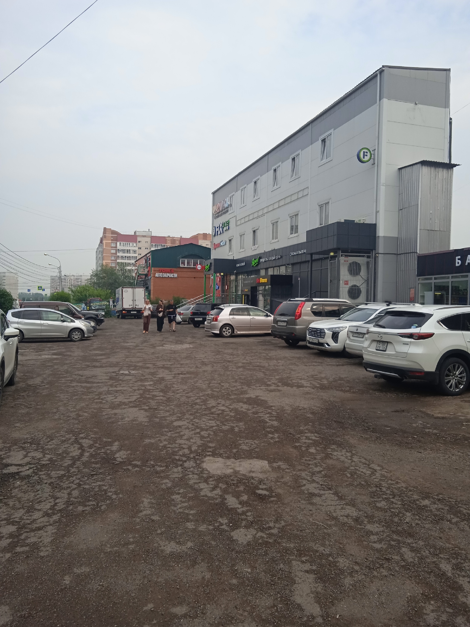 The image shows a parking lot in front of a modern commercial building with a grey and white facade. Several cars are parked on the lot, including a white SUV and a silver sedan in the foreground. Three women are walking across the parking lot towards the building. The building appears to be a retail or office space, with signs visible in Russian. In the background, there are other buildings and trees, suggesting an urban environment. The sky is overcast, and the overall impression is that of an ordinary day in a city. The building on the right has a sign with the letter "F" in a green circle. A sign on a brick building further back reads "Автозапчасти" which translates to "Auto Parts". The location is the city of Krasnoyarsk, Russia.