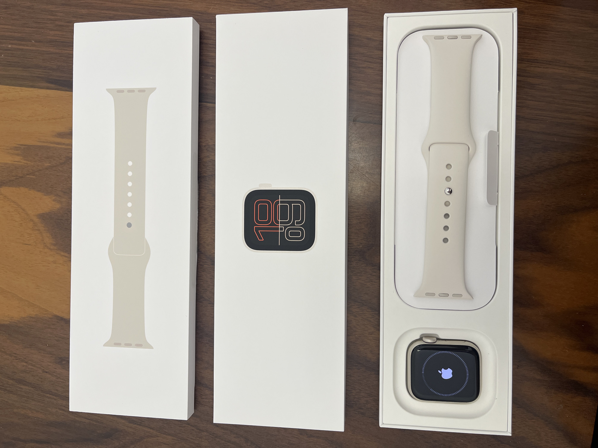The image shows three rectangular white boxes on a dark wooden surface. From left to right, the first box has a graphic of an Apple Watch band on it. The second box displays a stylized image of an Apple Watch screen, showing the time. The third box contains an Apple Watch with a charging screen and the band beside it. The setting appears to be indoors, likely a tabletop surface. The scene is devoid of human presence or action.