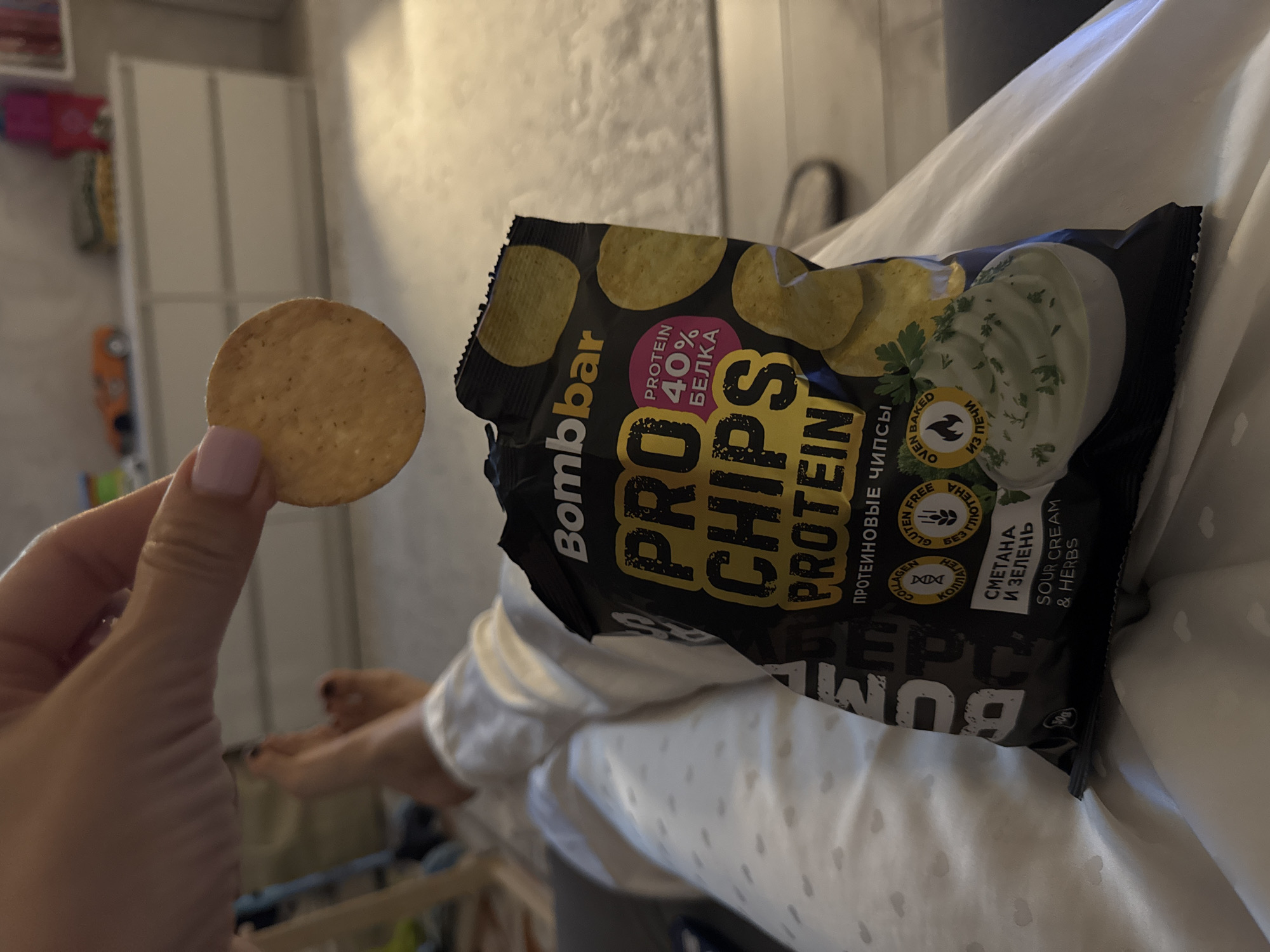 A close-up, indoor, medium shot captures a person's hand holding a single, circular, golden-brown cracker over a bag of "Bombbar PRO CHIPS PROTEIN" snacks. The bag, predominantly black with yellow and pink text, displays imagery of potato chips and a dollop of sour cream and herb dip. The background is blurred but suggests a domestic setting with furniture, including a white dresser. The person's legs, clad in white patterned pajamas, are visible in the lower half of the frame, indicating a relaxed, seated posture. The scene is not located in the city of Sochi, Russia.