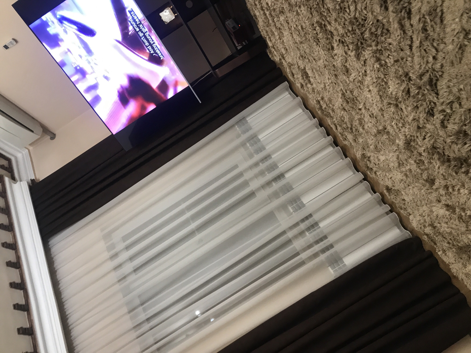 The image captures an interior setting, likely within a home, featuring a television displaying vibrant imagery, partially visible curtains, and a textured rug. The curtains have both sheer white and dark brown panels. The television is displaying an animated or stylized image. The environment is somewhat dimly lit. There's no definitive evidence to pinpoint the location as Yerevan, Armenia, or the presence of any people, actions, or interactions in this particular frame. The image is primarily focused on the physical elements within the room.