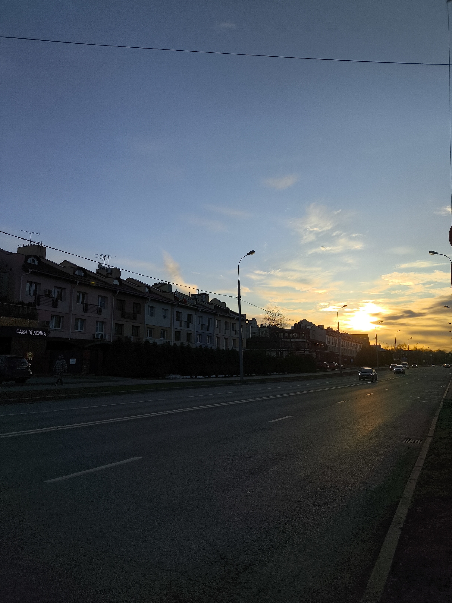 The image shows a street in Moscow, Russia, at sunset. The sky is a mix of blue and orange, with the sun low on the horizon. A row of buildings lines the left side of the street, including "CASA DI NONNA". A row of hedges sits between the building and the road. Several cars are driving on the road. Streetlights are visible along the sides of the road. A pedestrian is walking on the sidewalk.