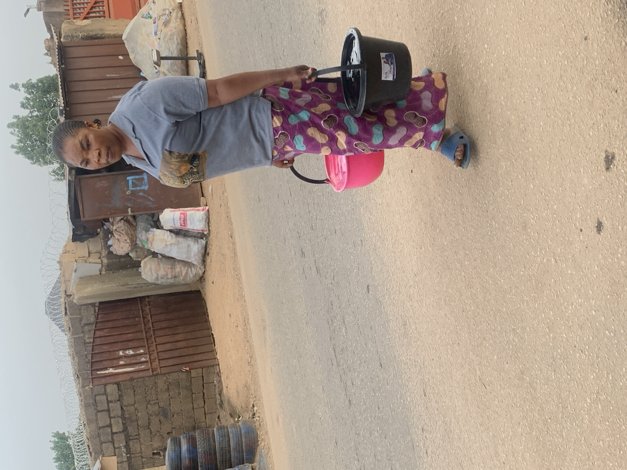 An adult woman stands on a paved road, carrying a black bucket in her right hand and a pink bucket in her left. She is wearing a gray shirt, patterned purple pants, and blue sandals. The background includes a brown metal gate, a cement block wall with barbed wire, and bags of what appears to be trash or recycling. Stacked car tires are visible near the bottom of the image. Given the visual context, the location is likely a residential street within the city of Jos, Nigeria.