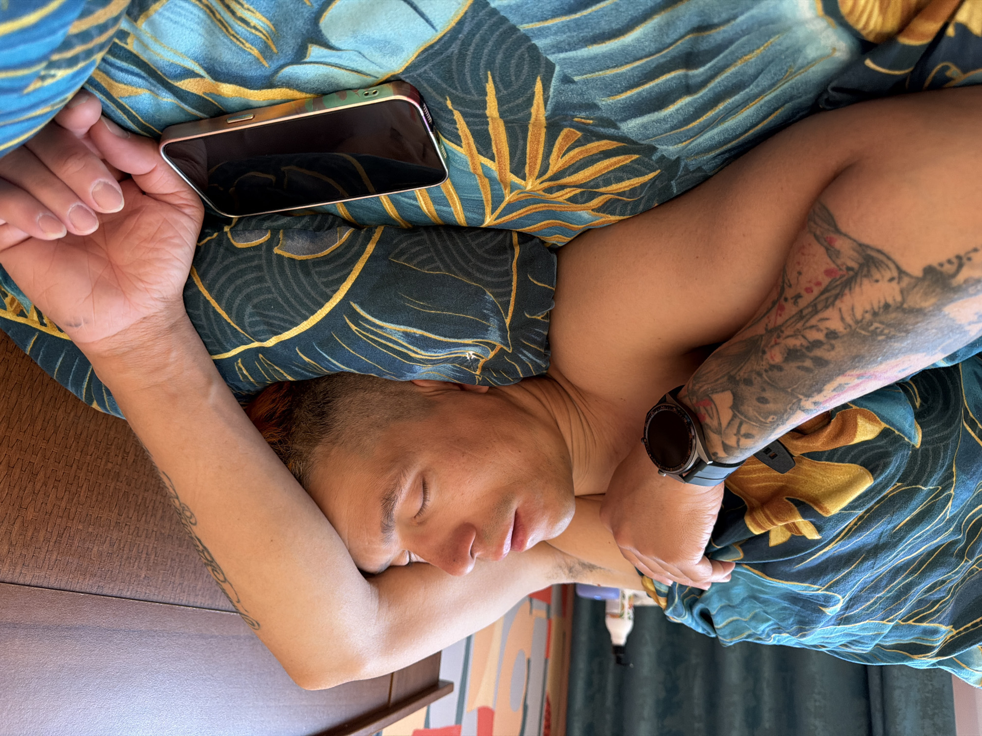 A man with a tattoo on his left arm lies asleep on a blue and gold patterned bedspread. His right arm rests on a dark wooden surface, and his phone lies next to his left hand. A watch is visible on his left wrist. The location is Chaikovsky, Russia.