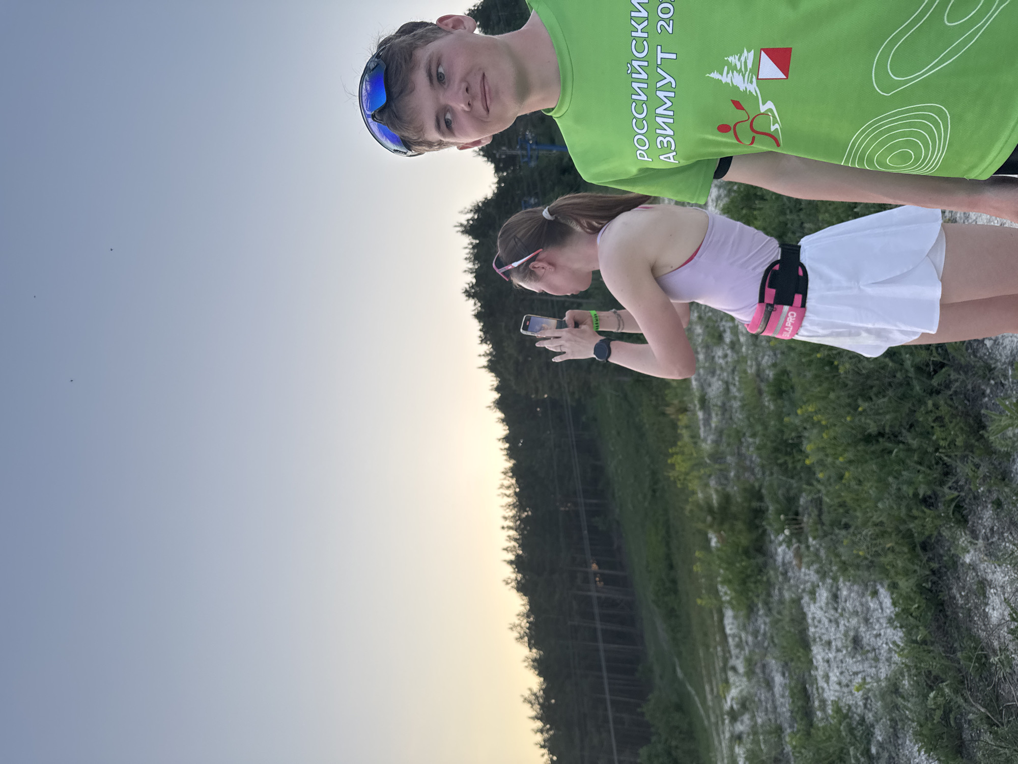 Two people are in the photo. A male with short brown hair is wearing a green t-shirt and sunglasses on top of his head. A female with long brown hair, in a ponytail, is wearing a pink tank top, white running skirt, and a hydration belt. She is holding a smartphone and appears to be taking a photo. The background includes a forest, sandy terrain, and a light-colored sky. The t-shirt worn by the male contains the text "РОССИЙСКИЙ АЗИМУТ 2021." They are likely participating in an orienteering event. It is daytime and the location may be Khvalynsk, Russia, based on the text of the shirt.