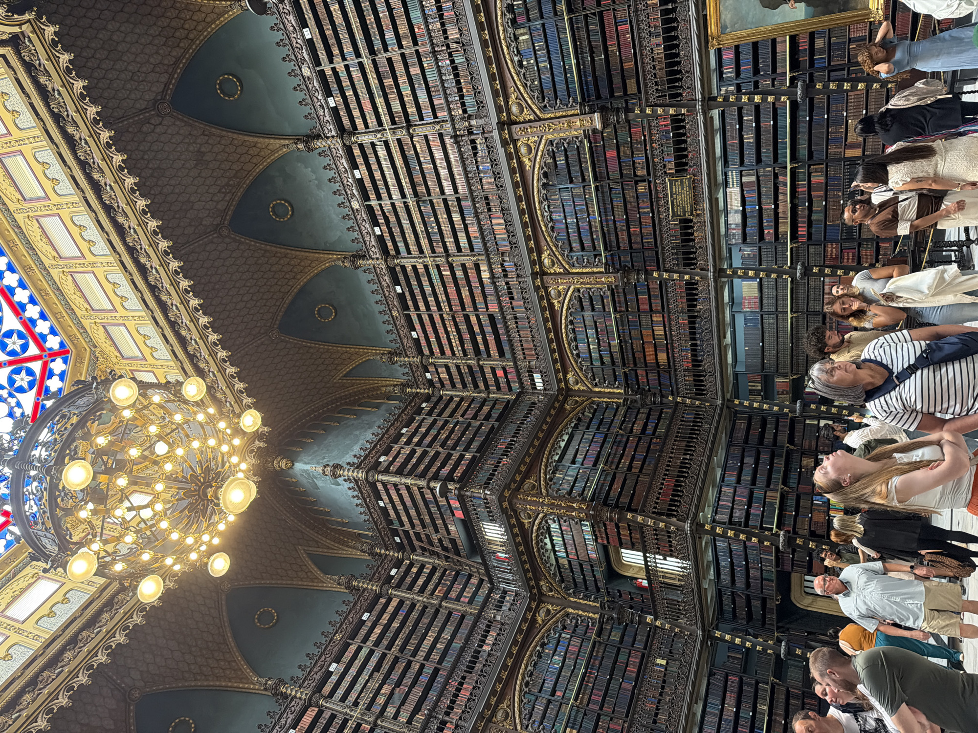 The image depicts the interior of the Real Gabinete Português de Leitura in Rio de Janeiro, Brazil. This historic library is characterized by its towering, ornate bookshelves filled with numerous books. A large, elaborate chandelier hangs from the ceiling, illuminating the space. The architecture features intricate detailing on the balconies and arches, with a stained-glass ceiling visible at the top. Several people are present in the foreground, some looking up at the architecture and books, while others are engaged in conversation. The atmosphere appears to be that of a tourist attraction or a public library visit.