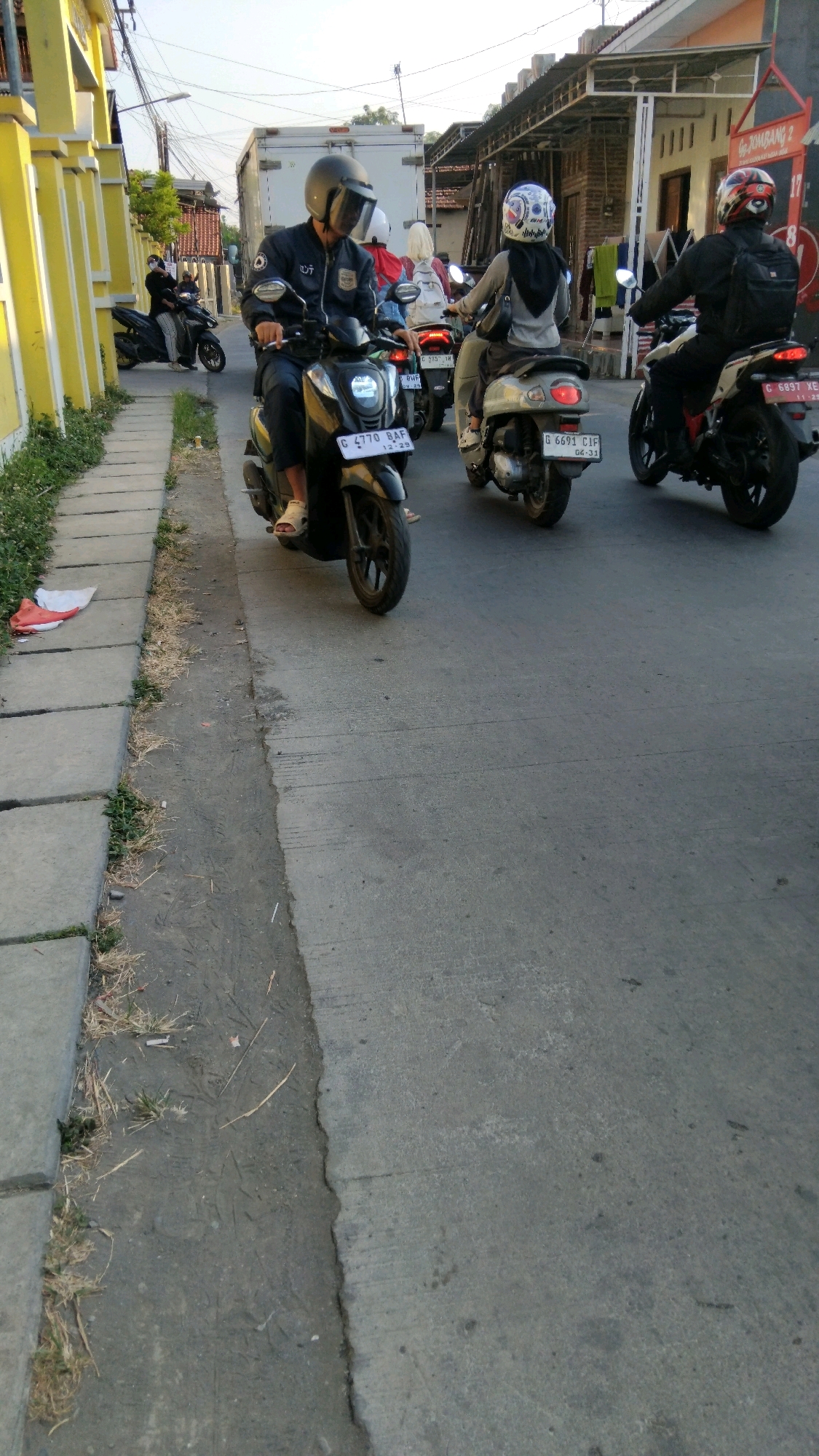 A group of motorcyclists are riding down a street in Tegal, Indonesia. The foreground is dominated by the grey asphalt road, with a concrete sidewalk on the left side of the frame. The sidewalk is lined with sparse green grass and dried vegetation. A large, red piece of plastic trash is lying on the sidewalk.
In the center of the image, a man wearing a dark jacket and a black helmet with a tinted visor is riding a black scooter. The scooter has a license plate that reads "G 4770 BAF". He is wearing white sandals. Behind him, a woman in a grey hoodie and a white and blue patterned helmet is riding a lighter-colored scooter. Her scooter's license plate is partially visible, reading "G 6691 CIF". Further in the background, a man wearing a black jacket and a red and white helmet is riding a white motorcycle. His motorcycle's license plate reads "C 6897 XE".
Other vehicles and buildings are visible in the background, including a white truck and houses with tiled roofs. The sky is a pale blue, suggesting it is daytime, possibly late afternoon due to the angle of the light. The overall scene depicts typical street traffic in an Indonesian city.