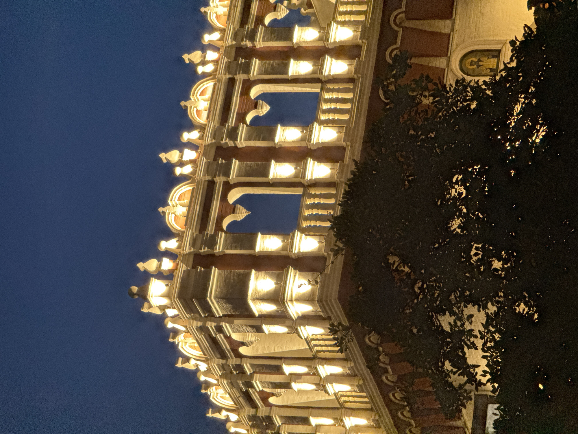 The image captures an illuminated section of the Spasskaya Tower in Moscow, Russia, at dusk. The architectural details of the tower are highlighted by warm, artificial lights, showcasing its red brick facade, white stone trim, and ornate decorative elements. Arched openings and decorative finials are visible along the upper levels. A dark silhouette of lush foliage from a tree partially obscures the lower portion of the tower. The sky above is a deep, clear blue, indicating the transition from day to night. The overall scene is one of historical grandeur illuminated against the evening sky.