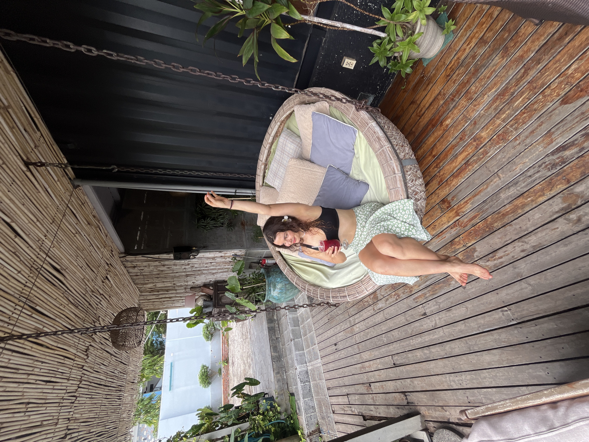 A woman is sitting in a wicker swing chair on a wooden deck in Mũi Né, Vietnam. The swing chair is adorned with several pillows in various shades of beige and gray, and a light green cushion. The woman is wearing a black bikini top and a light green skirt with a floral pattern. She is holding a glass of a dark red beverage with a straw in her right hand and has her left arm raised. A white flower is tucked behind her right ear. The deck is made of weathered wooden planks and is partially covered by a bamboo-like roof. To the left of the swing, there is a large potted plant. In the background, there is a dark corrugated metal wall and further to the left, a glimpse of a paved outdoor area with more plants and a light blue wall. A woven light fixture hangs from the bamboo roof above the swing.
