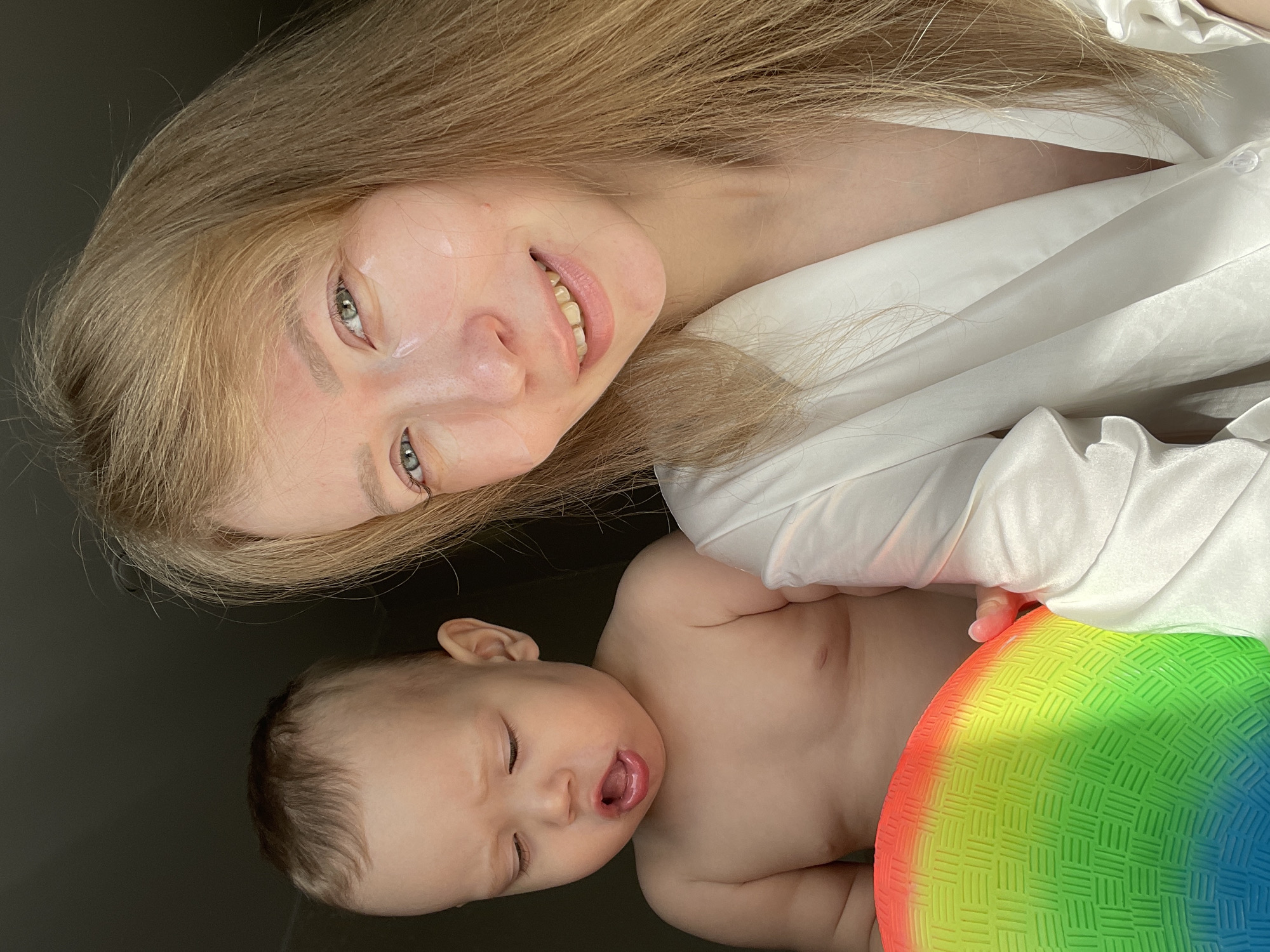 A portrait-style photograph features an adult and an infant. The adult, positioned at the top of the frame, has long, blonde hair and pale skin. She is wearing a white garment. The infant, located at the bottom, has fair skin and is shirtless. Beside the infant is a multi-colored ball, predominantly featuring hues of red, green, blue, and yellow. The background is a dark, neutral tone. This image was likely taken indoors, but a specific location or setting within Perm, Russia, cannot be definitively determined from this image alone.