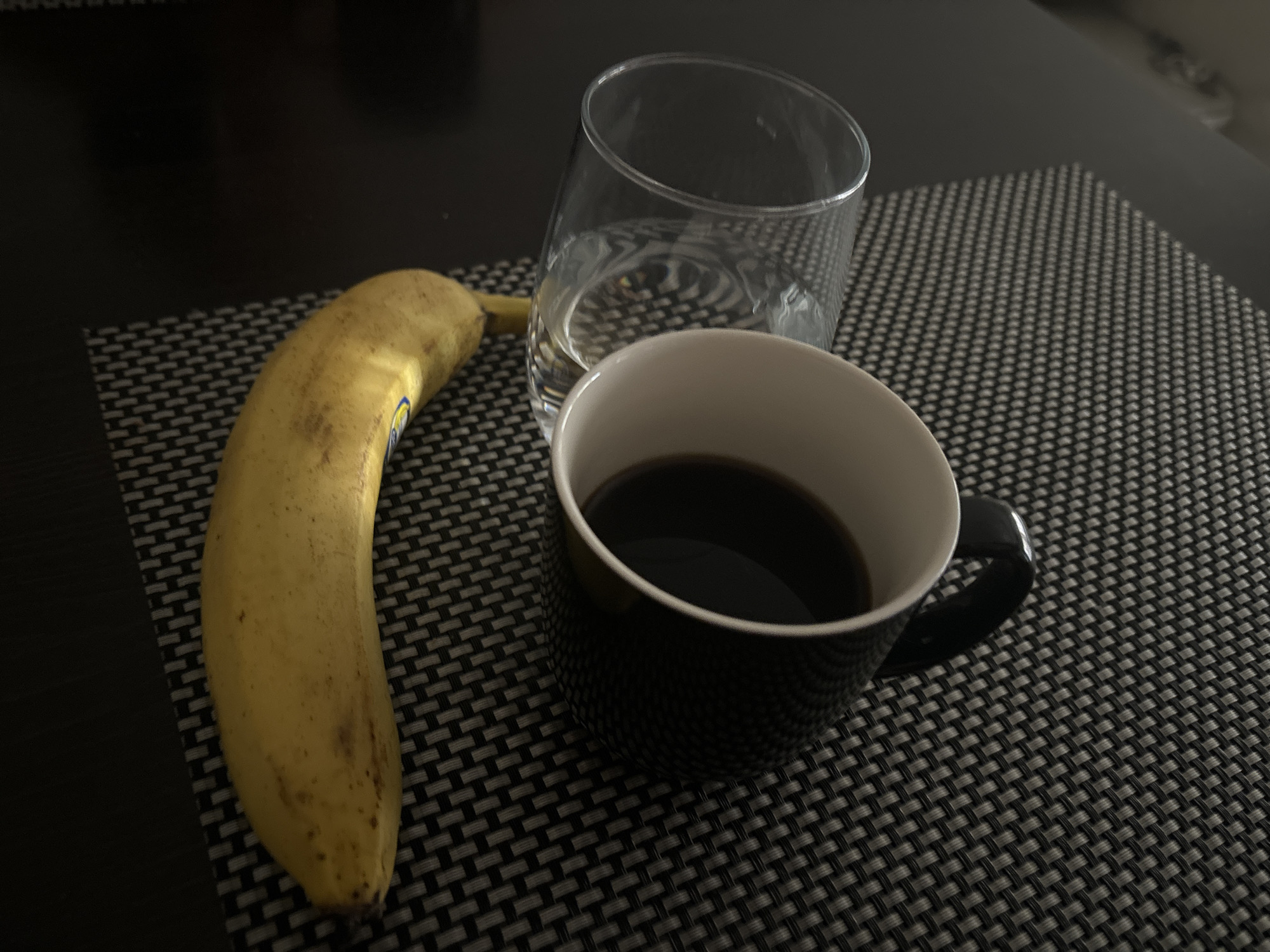 A banana, a glass of water, and a mug of coffee are on a black and gray woven placemat. The banana is yellow with some brown spots. The glass contains water. The mug is black with a white interior and contains coffee. The placemat is on a dark surface, possibly a table. The location is not discernible from the image.
