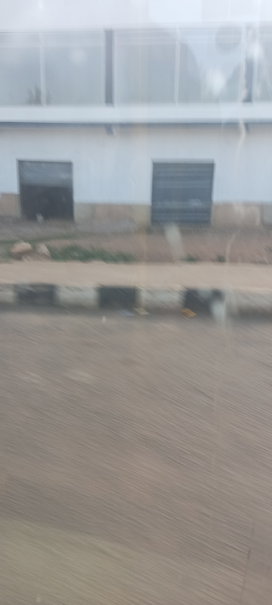 This blurry image, likely captured from a moving vehicle, depicts an outdoor scene in Gura Top, Nigeria. The foreground shows a road surface with prominent motion blur, indicating movement. A dark, block-like curb separates the road from an uneven patch of ground that features sparse green vegetation and some debris.
In the mid-ground, there is a building with a white lower wall. Two dark, rectangular openings, possibly windows with closed shutters or recessed doorways, are visible on this lower wall. Above the white wall, the upper section of the building appears lighter, possibly constructed with large, somewhat reflective or translucent panels, perhaps forming windows or a greenhouse-like structure, with faint vertical divisions.
Due to the significant blur, specific details are indistinct. No people or clear objects are identifiable beyond the architectural and landscape elements. There is no visible text. The scene appears to be taking place during the daytime, but the exact time or weather conditions are not discernible. The overall impression is one of passing by a building and roadside in an urban or semi-urban environment.