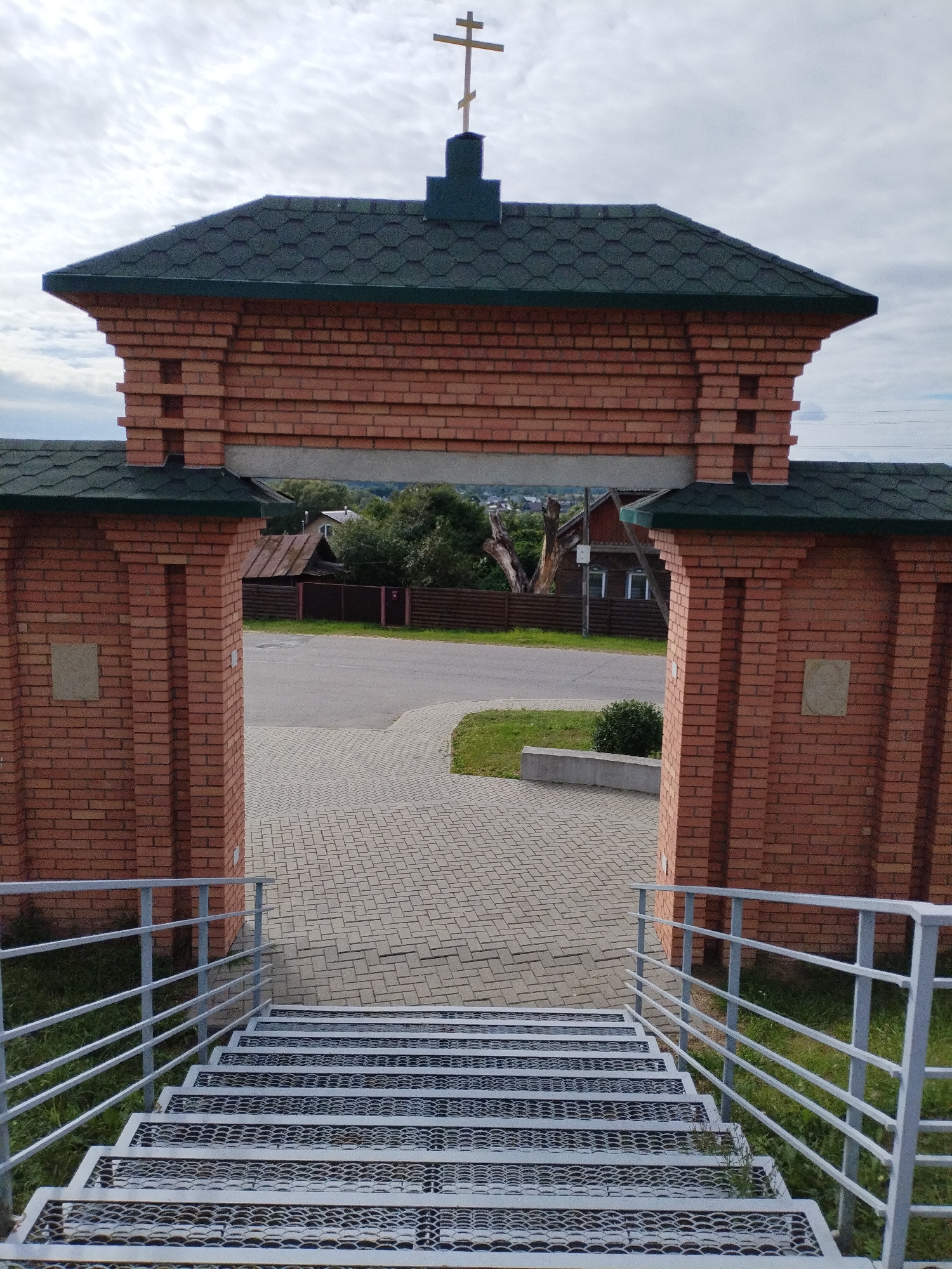 The image shows a view through a brick archway, looking out towards a village scene in Bialynichy, Belarus. In the foreground, a metal staircase with white railings descends from the viewer's perspective. The archway is constructed of red brick and topped with a dark green, shingled roof, capped with a wooden cross. Through the arch, a paved walkway leads to a road. Beyond the road, there are houses with wooden fences and trees. The sky is overcast with clouds.