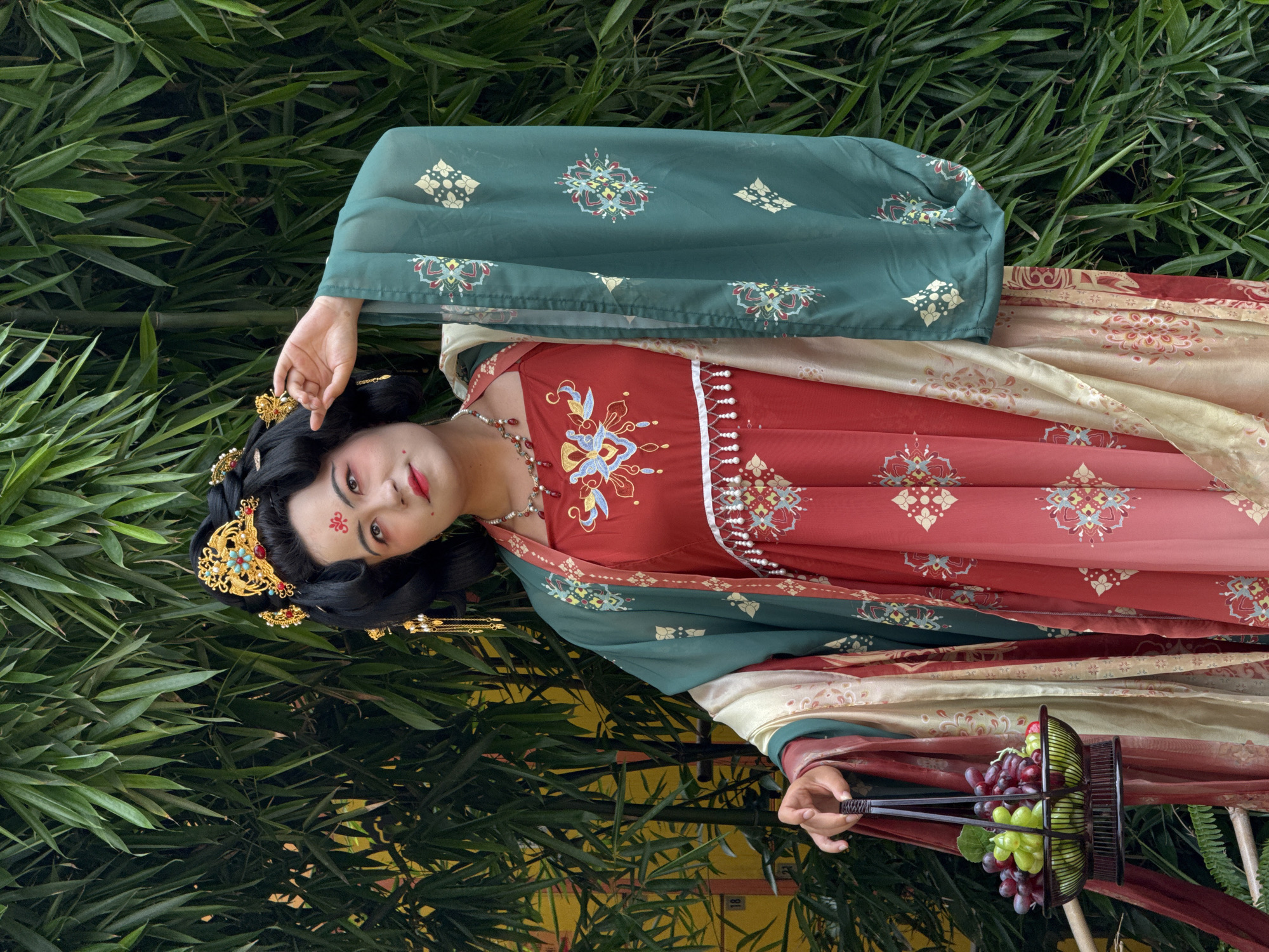 This portrait features a person in elaborate traditional Chinese attire, posing gracefully against a lush bamboo backdrop in Yuzhong District, China. The subject, likely female, has dark hair styled intricately with ornate gold hairpins adorned with red and turquoise jewels. A distinctive red huadian (forehead decoration) and elegant makeup, including winged eyeliner and red lipstick, complete the look.
She wears a multi-layered Hanfu-style outfit. The prominent outer layer is a dark teal-green fabric patterned with repeating motifs in gold, red, and blue. Beneath this, a vibrant red dress is richly embroidered with complex patterns in gold, blue, and white, and features pearl-like embellishments along its seams. A light cream or gold fabric with subtle reddish patterns forms an inner layer or sash. She also wears a silver-toned necklace with red beads.
In her right hand, she holds a black, open-weave basket filled with fresh red and green grapes. The background is dominated by dense, vibrant green bamboo leaves, suggesting an outdoor garden setting. A faint yellow structure with numbers '18' and '21' is partially visible behind the bamboo in the lower right. The natural, bright lighting indicates daytime. The overall image conveys a serene and artistic representation of traditional Chinese culture.