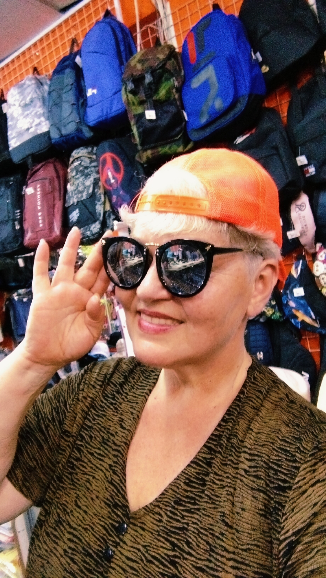 A woman with short, blonde hair wears an orange baseball cap and black sunglasses. She is smiling and making an "okay" hand gesture with her right hand. She is wearing a dark, patterned shirt. In the background, various backpacks are hanging on display in what appears to be a store. The scene is consistent with a retail environment in the city of Horishni Plavni, Ukraine.