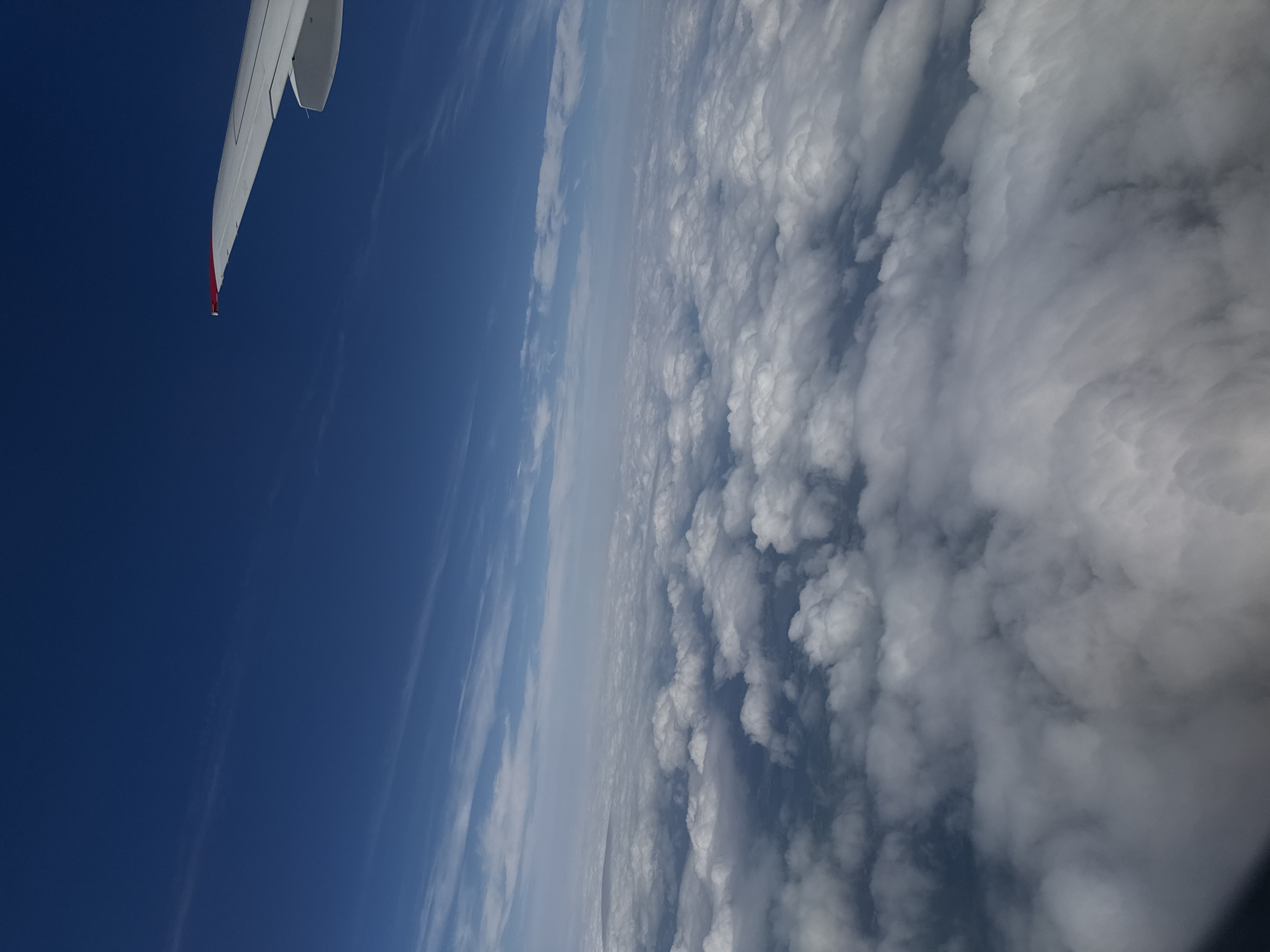 This is an aerial photograph taken from an airplane window, looking out over a vast expanse of clouds. The sky is a deep, clear blue, with thin, wispy clouds forming streaks across the upper portion of the image. Below, a thick blanket of white, puffy clouds stretches to the horizon, obscuring the ground. The wing of the airplane, painted white with a red tip, is visible in the upper right corner, extending into the blue sky. The scene conveys a sense of height and distance, with the clouds creating a soft, undulating landscape below. There is no visible information to indicate the location in Staroe Mart'yanovo, Russia.