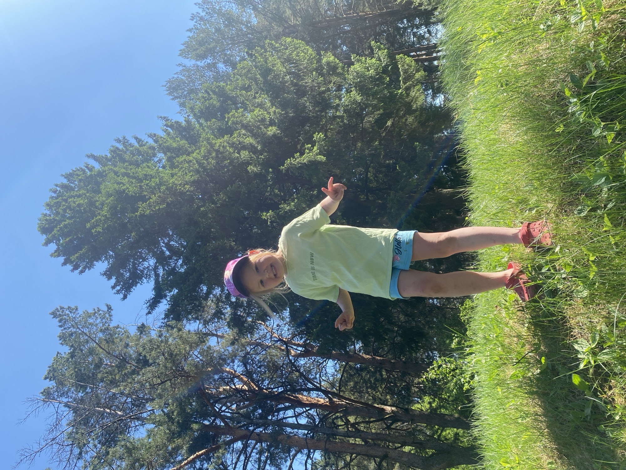 A young girl stands in a grassy field in what appears to be a park-like area on a sunny day, possibly in the city of Chaikovsky, Russia. She is wearing a light green t-shirt, blue shorts, a colorful cap, and red shoes. The girl is positioned with her legs on the grass and trees behind her. The sky is visible with blue in the upper corner. Tall trees frame the scene. The orientation of the image is landscape and slightly rotated. The girl has her hand up in a "rock on" gesture and has a friendly smile.