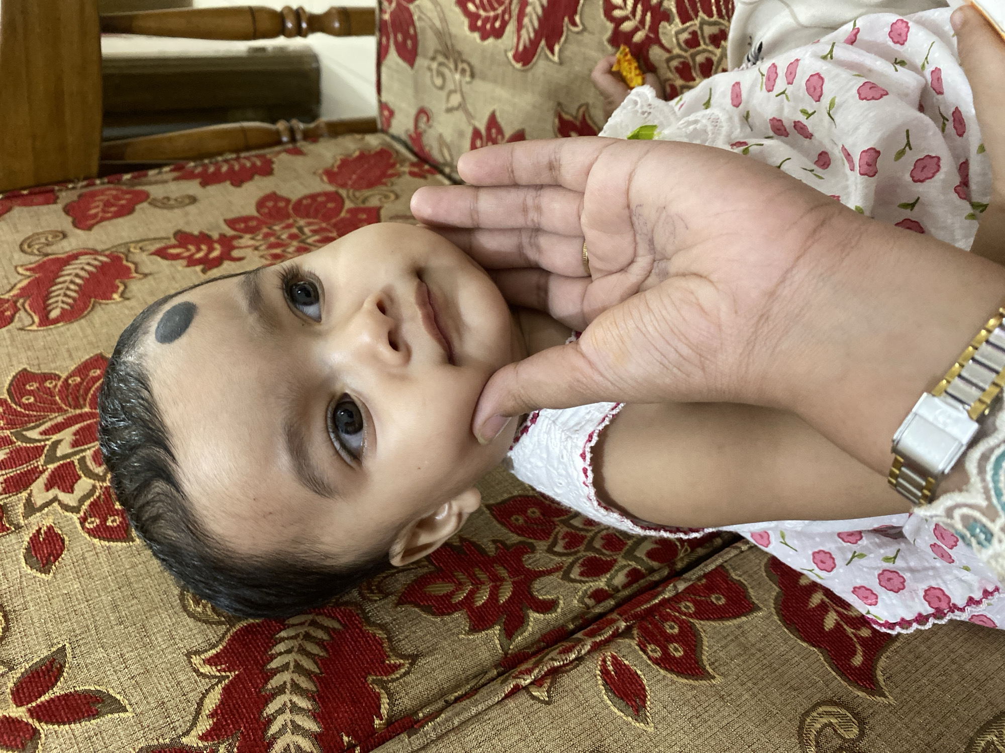 A baby lies on a patterned cushion, their face angled towards the camera. A hand with a wristwatch touches the baby's face. The baby has a black mark on their forehead. The surrounding environment suggests an indoor setting, potentially a home interior. There are no specific cues pointing to the city of Dhaka, Bangladesh.