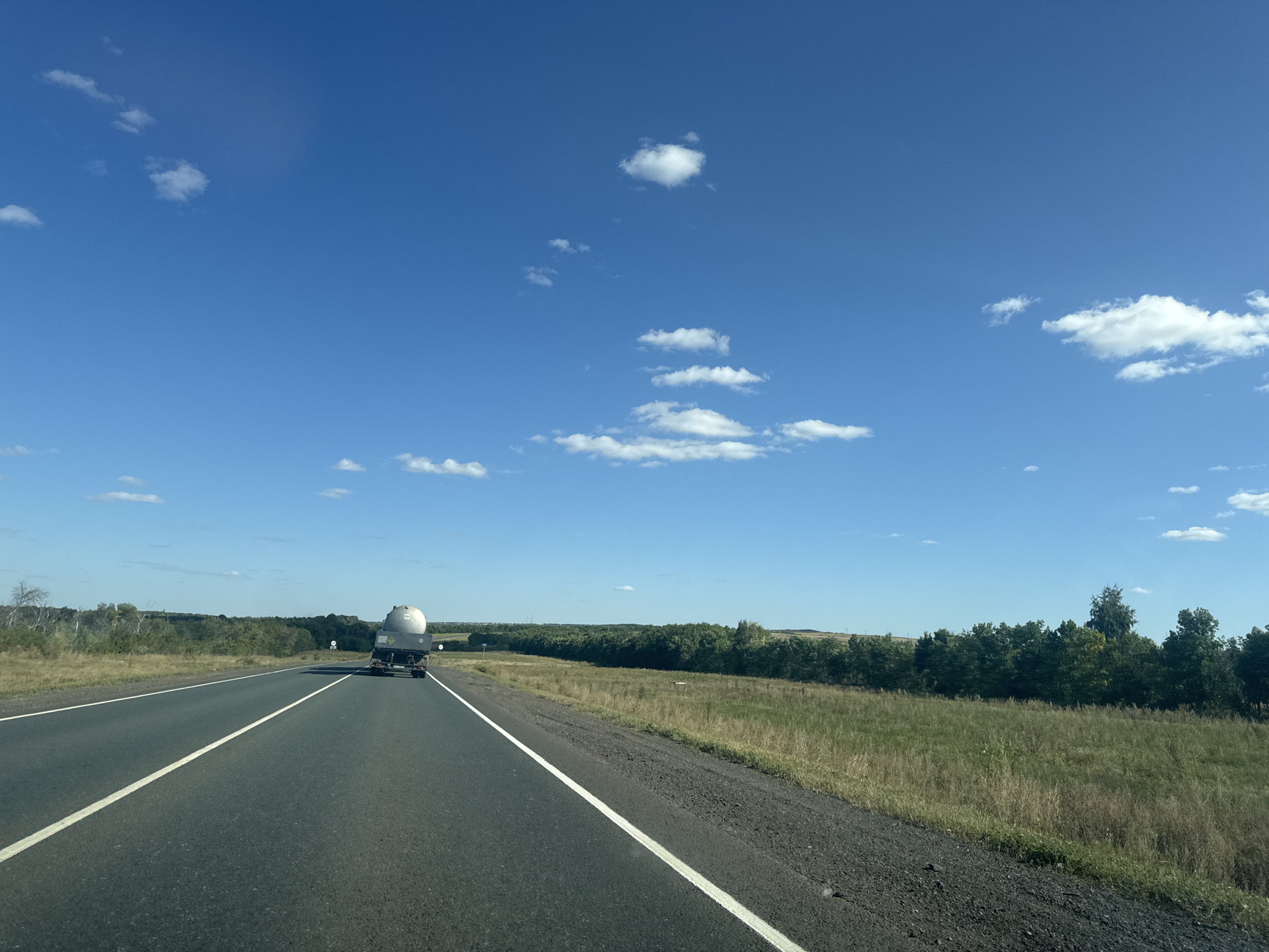 The image shows a view from a vehicle traveling on a paved road with white lane markings. The road curves slightly to the right. On the left side of the road, there is a dry grassy field leading to a line of trees in the distance. On the right side of the road, there is also a grassy field with scattered trees and shrubs, also extending to a line of trees in the distance. A large white tanker truck is visible driving ahead on the road. The sky is bright blue with scattered white clouds. The scene is consistent with a rural highway in the countryside of Perevolotsky, Russia.