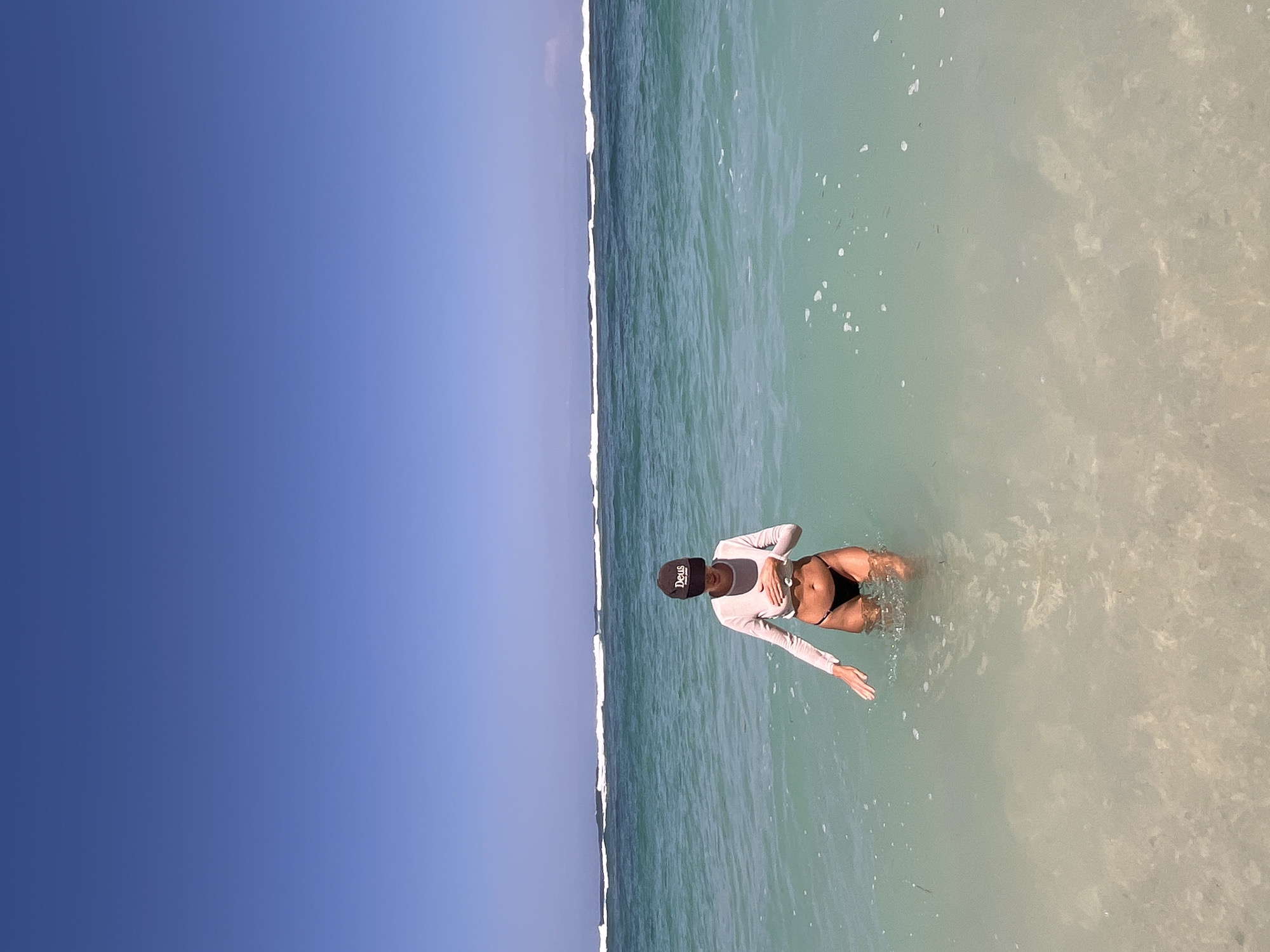 A woman stands in shallow, clear turquoise water at a beach in Kuta Selatan, Indonesia. She is wearing a long-sleeved light pink top and black bikini bottoms. A dark baseball cap with white lettering is on her head. The woman's left hand is on her chest, and her right hand is extended outwards, submerged in the water. Gentle waves with white foam are visible further out in the ocean, and the sky is a clear, bright blue. The sandy bottom is visible through the transparent water.