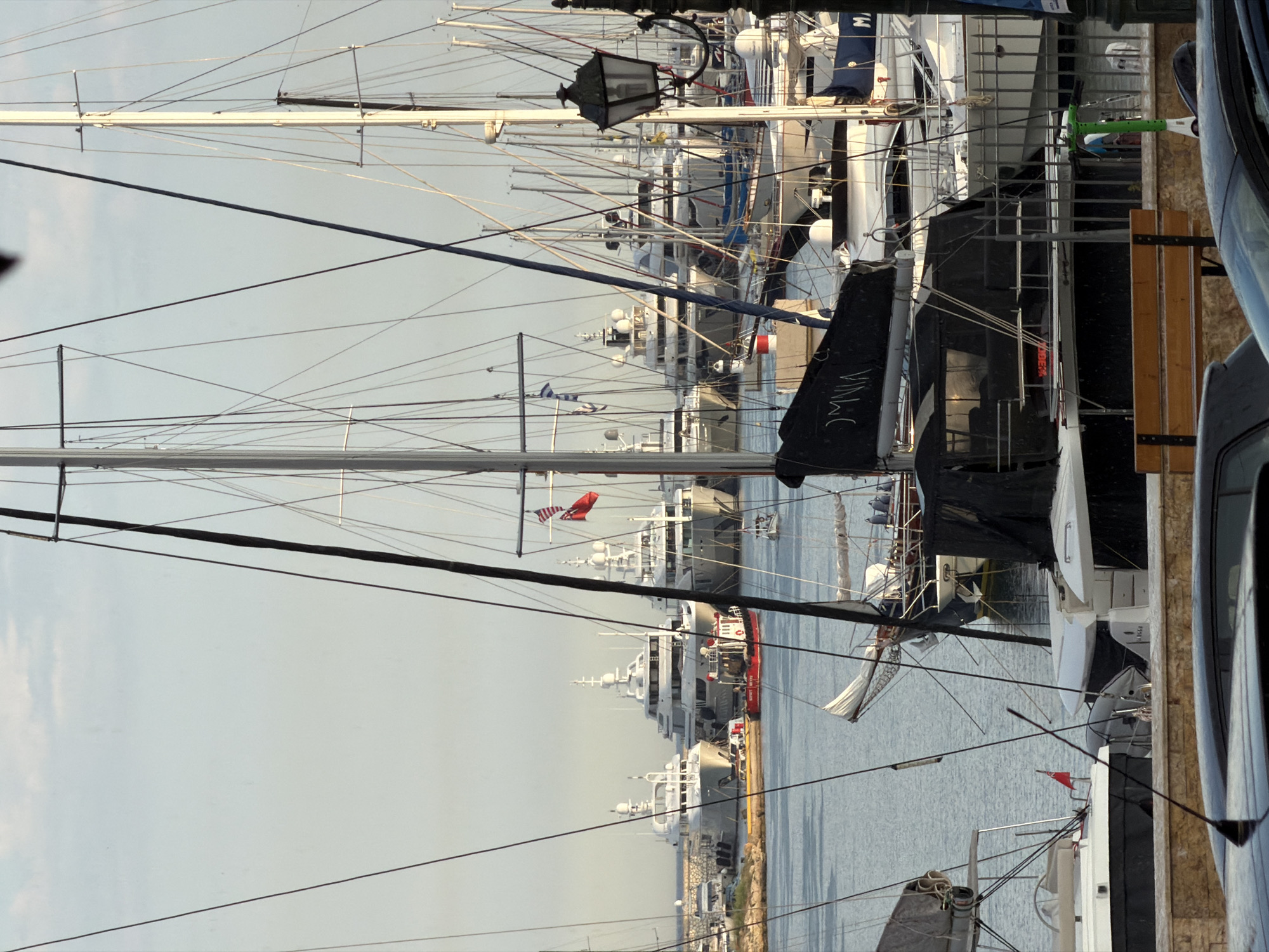 The image captures a busy harbor scene in Piraeus, Greece. Dominating the foreground are several yachts and sailboats docked side-by-side. The masts create a dense web of lines reaching upwards. One yacht features a dark sail cover labeled "VINA C." A bench with wooden slats sits near the edge of the dock. To the right of the frame, parts of vehicles are visible, suggesting a road or parking area adjacent to the harbor. In the background, larger motor yachts and cruise ships are docked, contributing to the crowded port. A few national flags are visible on some of the boats. The water is calm, reflecting the gray sky above. The scene shows a mix of leisure and commercial maritime activity.