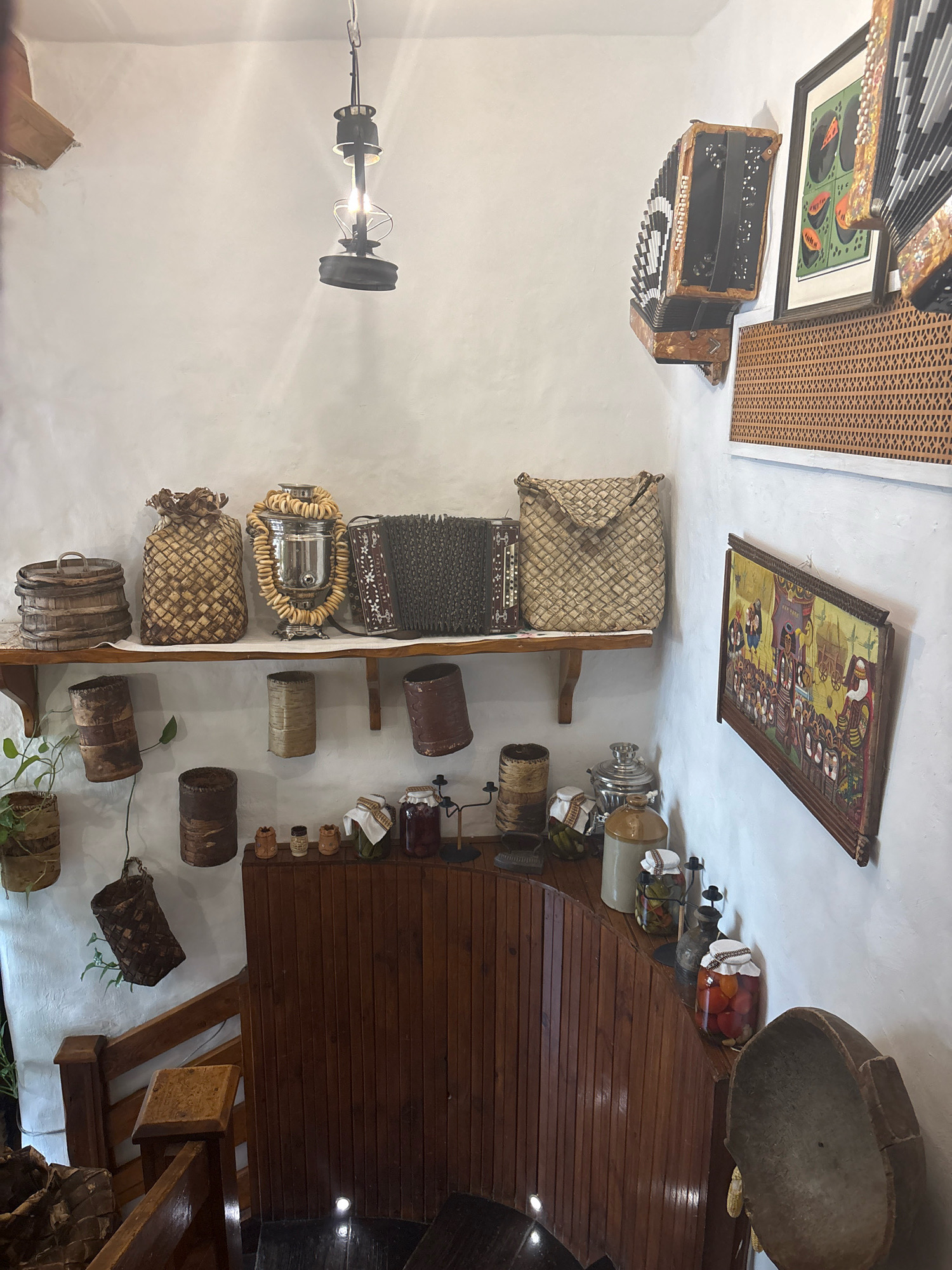 This image shows an interior scene, likely a museum exhibit or a rustic themed shop, with an abundance of traditional folk art and items, reminiscent of Russian village life. On the upper shelf, a variety of woven baskets, a samovar adorned with bagels, and an accordion are displayed against a white textured wall. Below this shelf, several cylindrical wooden and bark containers hang, with a potted plant nearby. The lower part of the image features a collection of jars filled with preserved food items, such as pickles and tomatoes, and a few smaller wooden decorative items on a wooden counter. To the right, a framed folk art painting depicts a procession, and another accordion is mounted on the wall. The overall impression is one of rustic charm and a celebration of traditional crafts. The scene does not provide any specific visual cues to pinpoint its location within Moscow, Russia, but rather conveys a generalized sense of Russian heritage. There are no people present in the image, and no actions are being depicted. The environment is indoors, with a warm and inviting atmosphere created by the wooden elements and the displayed artifacts.