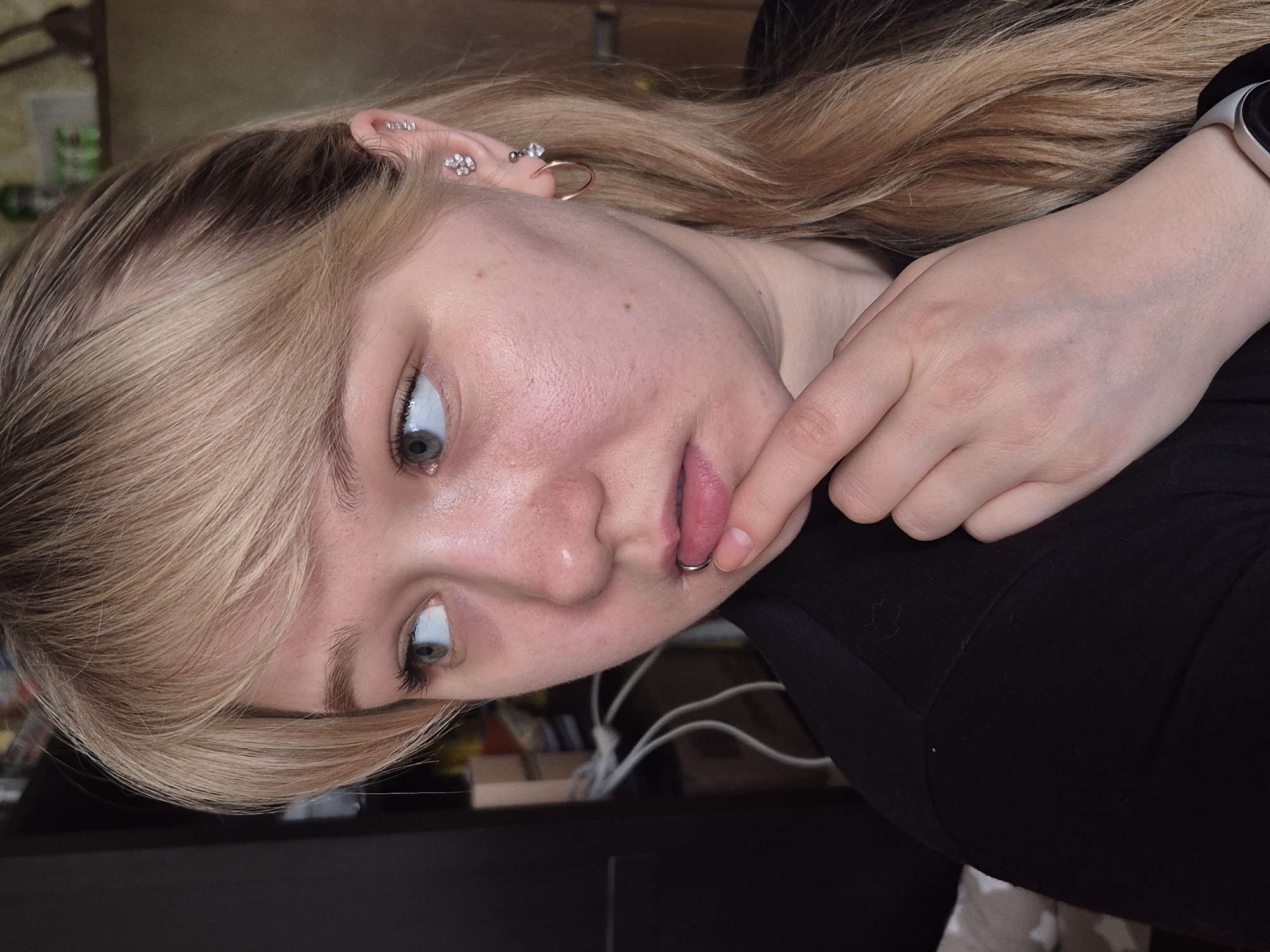 A young woman with light blonde hair and blue eyes is positioned in the foreground of the image. She is wearing a black top and has multiple piercings in her left ear, including studs and a hoop earring. She also has a piercing on her lower lip, which she is touching with her right index finger. A smartwatch is visible on her right wrist.
The background is blurry and contains indistinct objects. Based on visual cues, it is not possible to confirm with certainty that the image was taken in Chaikovsky, Russia.