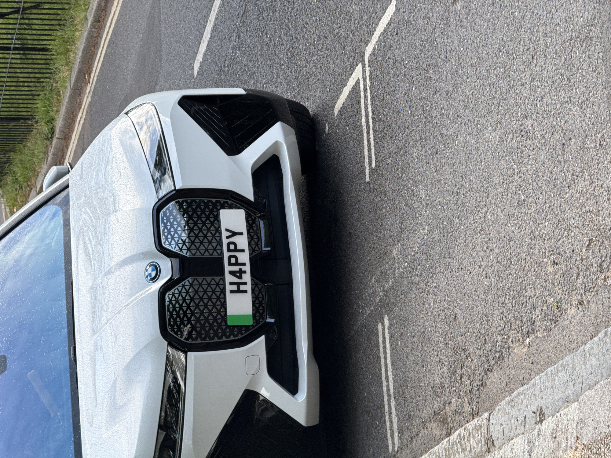 A white BMW with a personalized license plate that reads "H4PPY" is parked on a street in London, United Kingdom. The car is positioned close to the curb and oriented at an angle relative to the viewer. The front of the car is visible, showcasing its kidney grille and the BMW logo. The background includes a road with painted lines, a sidewalk, and a black metal fence. The scene is outdoors and the lighting suggests it is daytime. There are water droplets on the car's windshield.