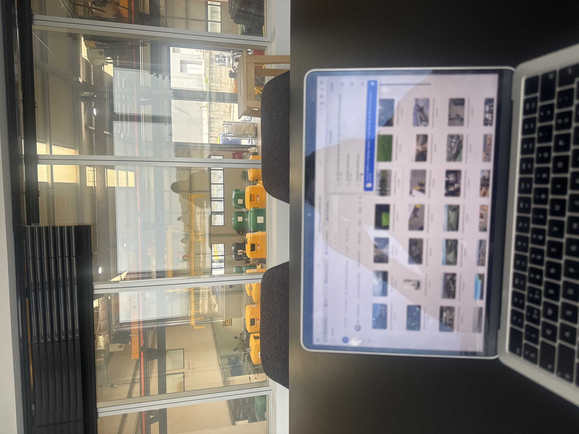 The image, rotated 90 degrees clockwise, presents an indoor scene looking out through a large window into an industrial or workshop area, likely in Torres Vedras, Portugal. In the foreground, a dark grey desk or table is visible, on which an open laptop rests. The laptop screen displays a gallery-like view of numerous small images or files, with a blue scrollbar on the right. The silver keyboard of the laptop is partially visible on the right side of the frame.
Beyond the desk, two dark grey chairs with upholstered backs are positioned, facing the window. The window itself is framed in white, comprising multiple large panes of glass. Through these windows, the interior of what appears to be a facility or external yard is visible. This area contains several industrial-style containers or pieces of equipment, predominantly yellow and green. One yellow container clearly shows the label "P5", and a green one appears to have text like "RECICLE" (Portuguese for recycle). The reflections on the glass show structural elements from both inside and outside the building.
The background, seen through the window, includes industrial buildings with corrugated siding and other structures. The natural light suggests it is daytime. No people are visible, and the scene depicts a static view of a functional environment, possibly an office or viewing area within an industrial setting.