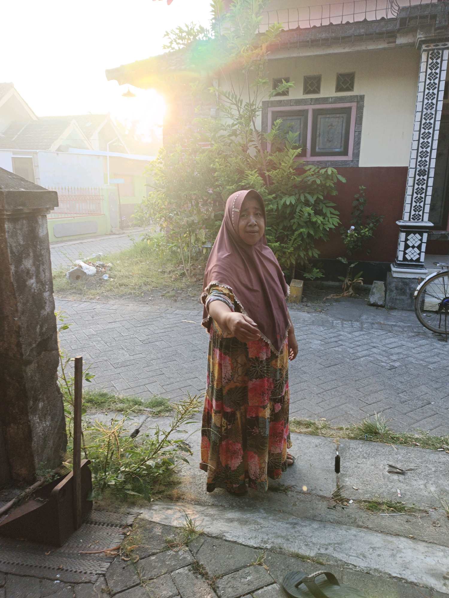 In Ketegan, Indonesia, an elderly woman wearing a maroon hijab and a long, colorful floral dress stands in what appears to be the entryway of a modest home. She extends her right hand forward, palm open, as if offering a greeting or invitation. The background reveals a house with white and dark red walls, adorned with decorative window grills and a black and white patterned pillar. Sunlight streams from the upper left, creating lens flare and illuminating the scene with a warm glow. The ground is paved with interlocking grey bricks, and patches of green grass and foliage grow along the pathway. To the right, a portion of a bicycle wheel is visible. In the foreground, a rusty dustpan rests against a textured stone pillar on the left. The overall impression is a candid, everyday moment captured in a residential setting.