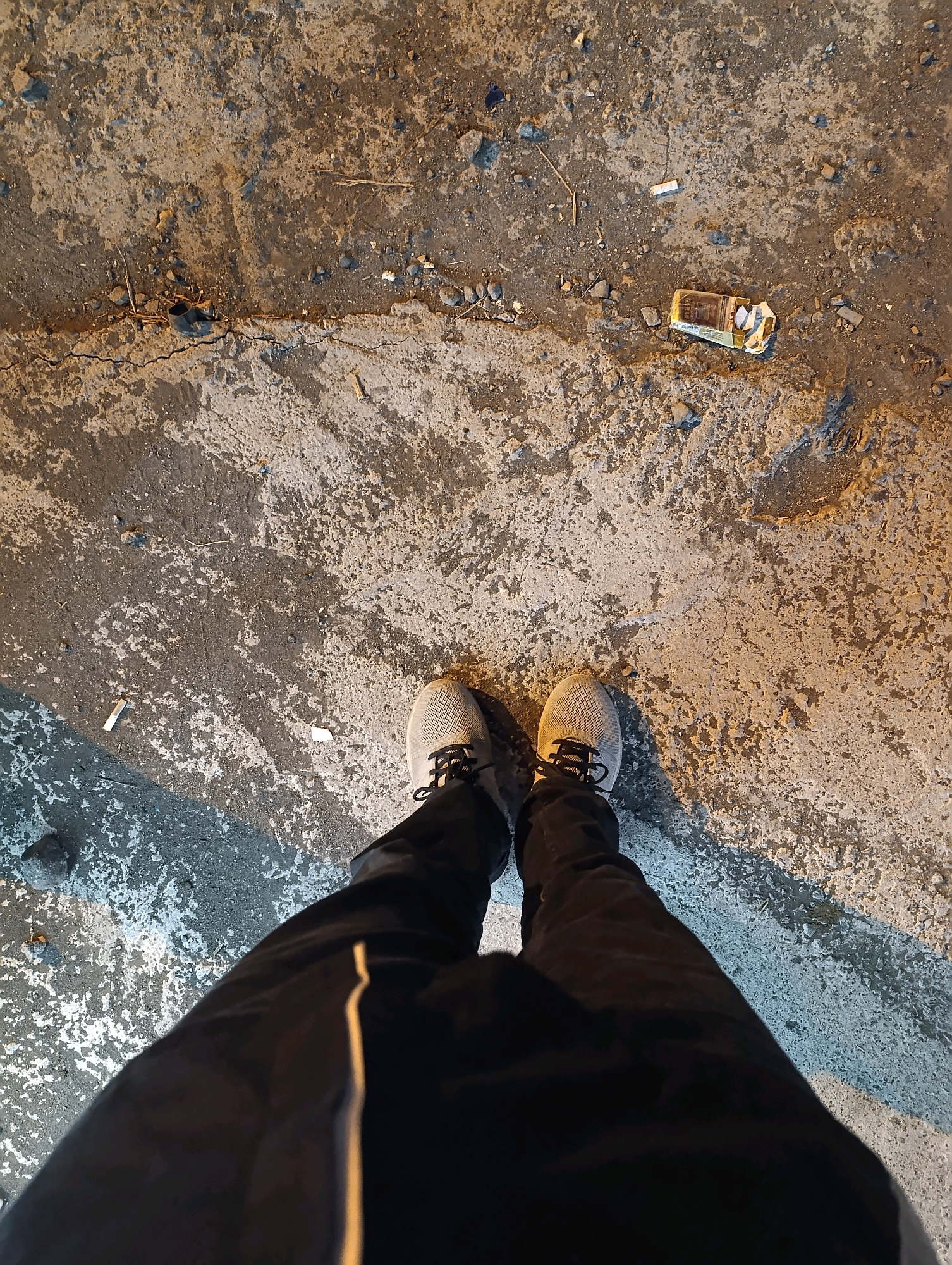 An overhead, eye-level shot shows a pair of light gray athletic shoes with black laces, standing on a cracked concrete surface. The shoes are worn by the subject whose dark pants, with a thin yellow stripe down the side of the left leg, are visible from the knees down. The concrete is strewn with small pebbles, dirt, and a crumpled cigarette pack with several cigarette butts scattered around it. A faint shadow falls across the concrete from the right, suggesting artificial lighting. The overall impression is a gritty urban environment, possibly a sidewalk or street in the city of Karangsono, Indonesia, where litter is visible.