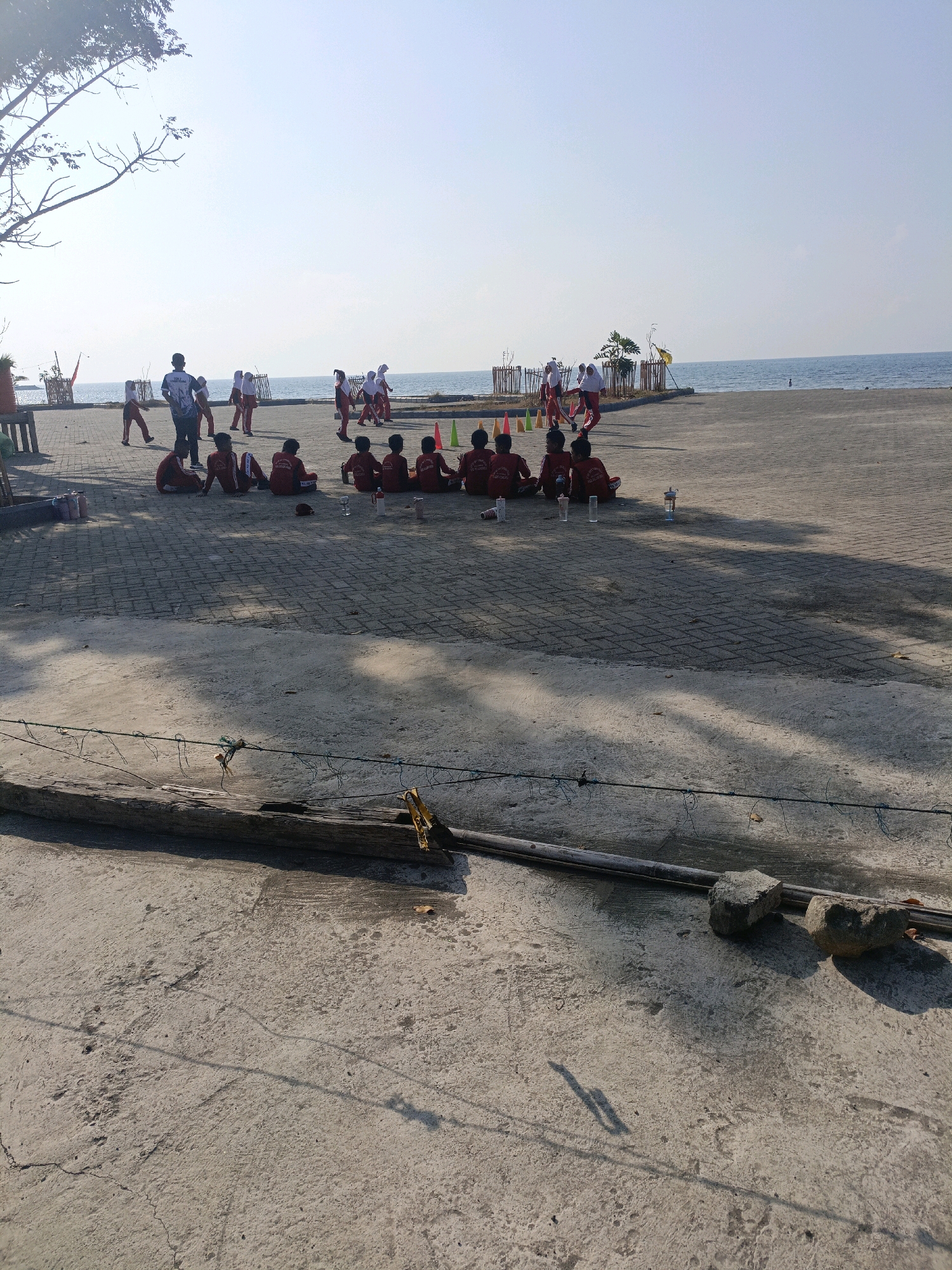 This is a medium-shot, outdoor photograph taken on a sunny day, likely at a beachfront location in Bulukumba, Indonesia. In the background, the blue ocean stretches to the horizon under a clear sky. The foreground is a paved area with gray interlocking bricks, casting long shadows from an unseen tree on the left.
In the middle ground, a group of children in red shirts and white headscarves are sitting on the paved ground, facing away from the camera. Some of them have water bottles in front of them. To the left of this seated group, a man in a dark shirt and pants stands, observing a group of children in similar red attire who are running and playing on the paved area near the water. Several orange traffic cones are visible in this area.
The immediate foreground consists of a concrete surface, cracked and weathered, with a few leaves scattered on it. A piece of weathered wood lies horizontally across the concrete, with a thin, taut wire stretched above it, possibly a makeshift barrier or clothesline. Two stones rest on the concrete near the right edge of the frame.
The overall scene depicts an outdoor activity session, possibly a school excursion or sports practice, near the coast. The presence of the ocean, the paved waterfront area, and the children engaged in activities suggest a public recreational space.