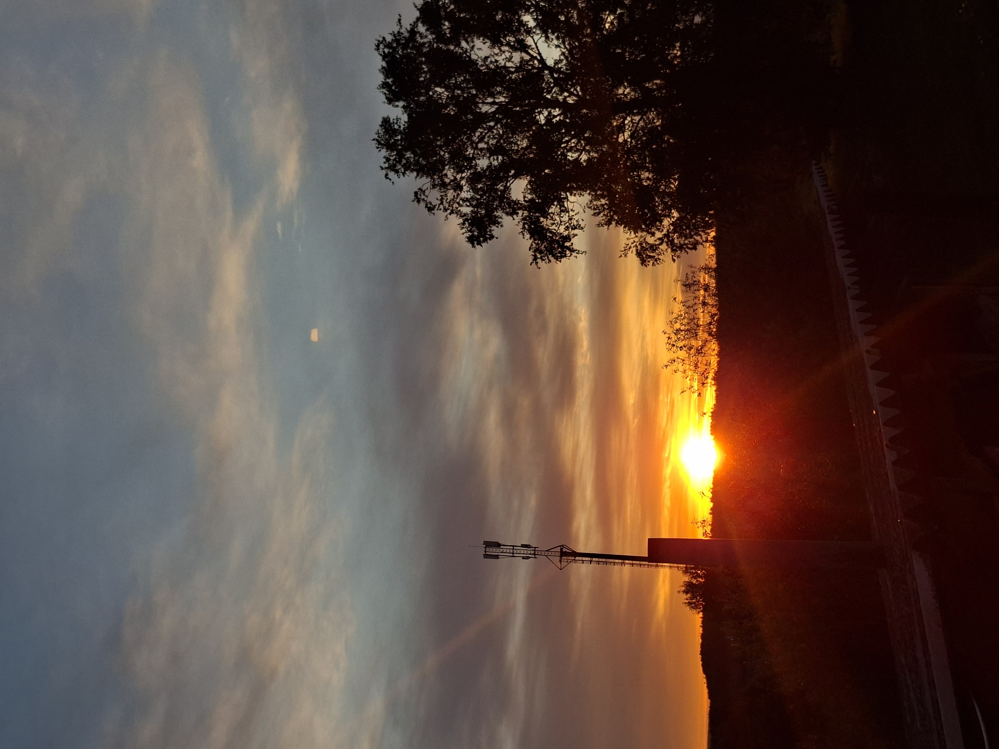 A vertical outdoor shot captures a vibrant sunset over a landscape. The sky transitions from blue-gray clouds on the left to orange hues near the center, where the sun is setting. A tree silhouette is visible at the top right. A tower structure is near the center-left. On the right, the landscape is marked by a dark, horizontal line, with the horizon ablaze with the sunset's glow. The location is Unknown, Unknown.