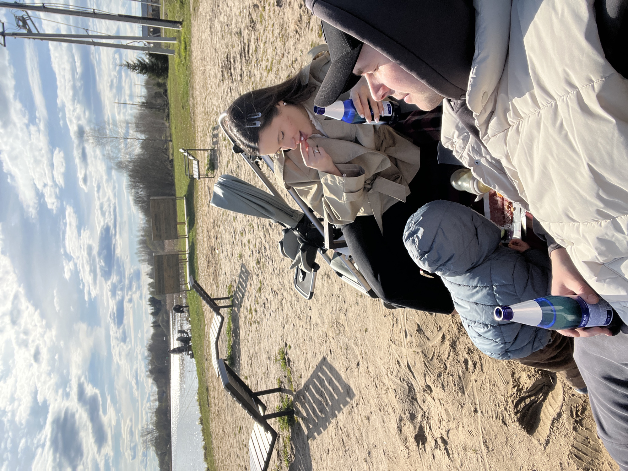 Observed is a scene outdoors, apparently located near a lake or a body of water, potentially within Литвиново, Russia. The image features three individuals: a woman eating a red fruit, a man next to her, and a child holding a bottle. The woman is seated near a baby stroller. The environment is characterized by a sandy terrain, benches, and a landscape that includes grass and trees. The sky is predominantly blue with white clouds.