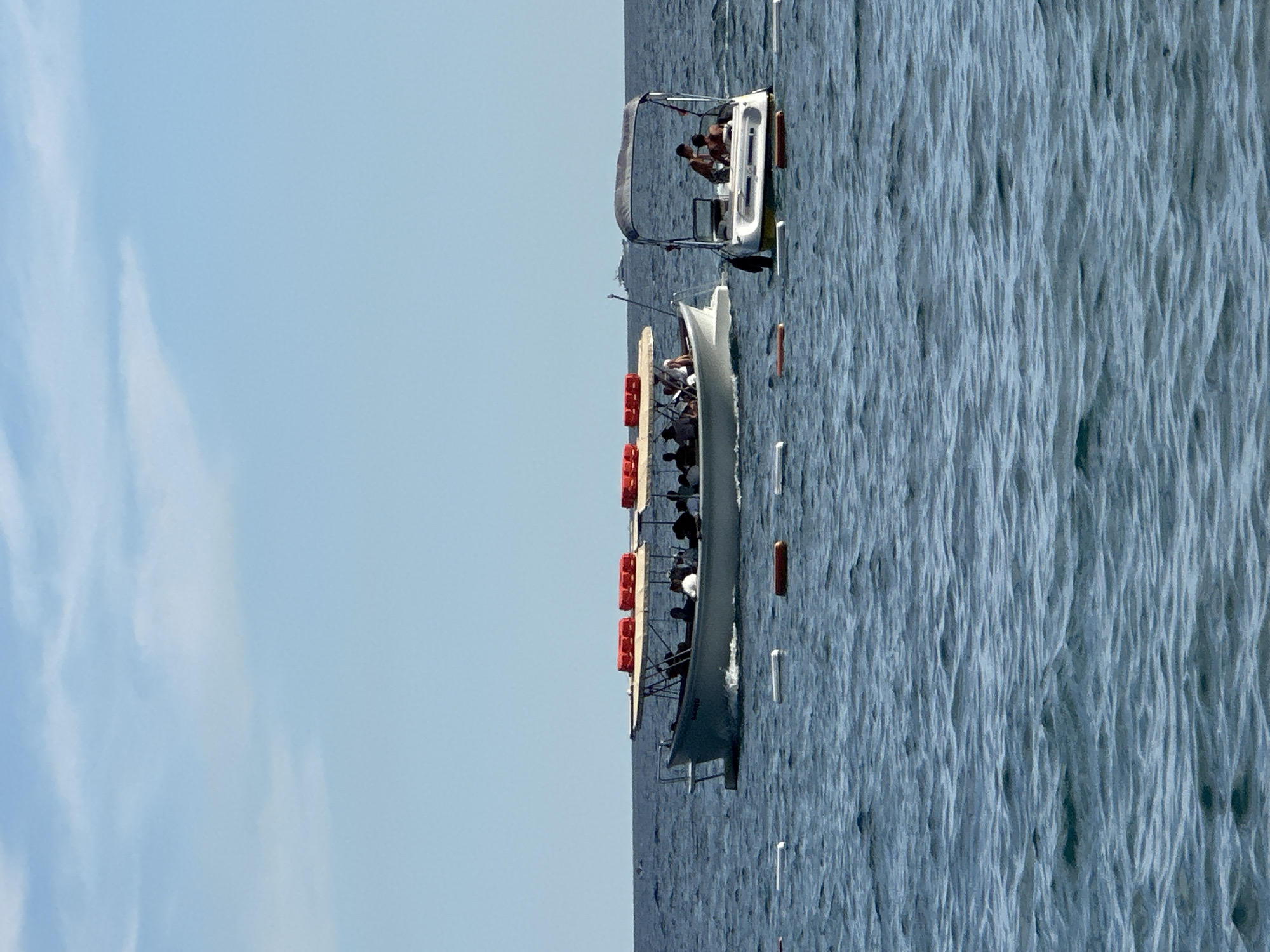 In the city of Boreti, Montenegro, two boats are visible on a body of water under a clear blue sky with scattered clouds. The boat on the left is larger and appears to be a tour boat, carrying multiple passengers under a shaded canopy. Orange life preservers are lined up on the canopy's edge. The boat on the right is smaller and has a canopy for shade. Three individuals are seated in this boat. Both boats are moving through the water, creating wakes. In the foreground, a series of white and orange buoys are floating on the water's surface, possibly marking a swimming area or boundary. The horizon line where the water meets the sky is visible.