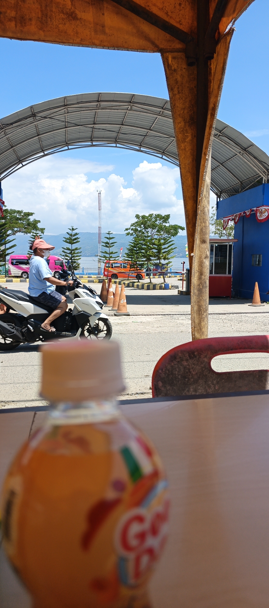 The image depicts a scene at a ferry terminal in Tigaras, Indonesia. In the foreground, a blurred orange bottle with a cap is positioned on a brown surface, partially obscuring the view of the rest of the scene. In the midground, a person wearing a light blue shirt, dark shorts, a pink cap, and a white face mask is riding a white scooter. They are moving from left to right across the frame. The scooter has a black seat and black wheels. Behind the scooter rider, a row of orange traffic cones is visible on the asphalt road. Further in the background, a bright blue building with red trim and a white kiosk are present. Red and white banners are draped across the blue building. A large, curved metal roof structure is visible, supported by an orange, weathered beam in the right foreground. Beyond the terminal, a body of water, likely Lake Toba, is visible, with hills and mountains in the distance under a bright blue sky with scattered clouds. Several evergreen trees are planted in the area. Two vehicles, a pink and white van and an orange van, are parked or moving in the background. The overall environment suggests a sunny day at a bustling transportation hub.