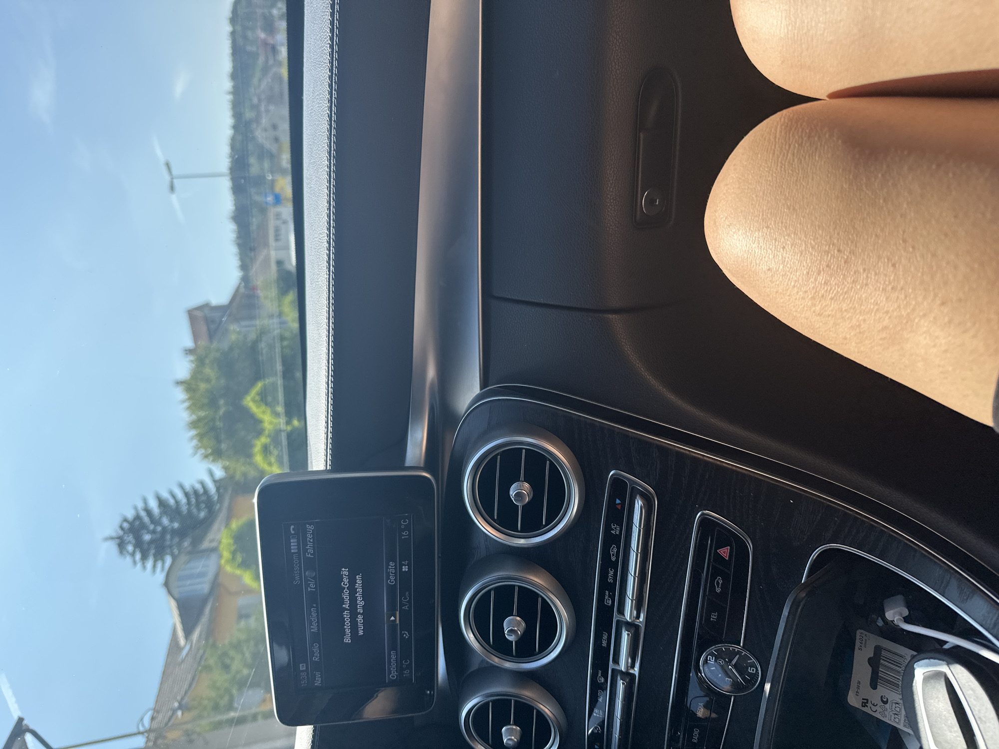 The image shows the interior of a Mercedes-Benz C-Class car on a sunny day, with the location identified as Räterschen, Switzerland. The car's dashboard is visible, featuring a central infotainment screen displaying "Bluetooth Audio-Gerät wurde angehalten." Below the screen are climate control vents and buttons. The digital clock on the dashboard reads 15:38. The view outside the car through the windshield shows a suburban area with houses and trees under a clear blue sky. The legs of a person are visible in the foreground, suggesting the photographer is a passenger. The overall scene is static and focused on the car's interior and immediate surroundings.