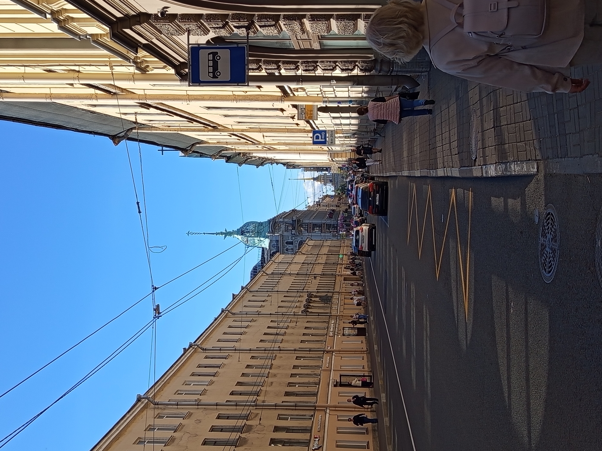This is a photograph of a street scene in Saint Petersburg, Russia, likely taken on Nevsky Prospekt, a major thoroughfare. The image shows a wide street with buildings lining both sides. On the left, a long, pale yellow building with numerous windows stretches into the distance. On the right, a cream-colored building with ornate architectural details and a dark mansard roof dominates the foreground.
The street is paved with asphalt and marked with yellow lines, indicating a pedestrian crossing. Several cars are visible on the road, some driving and others parked. People are walking on the sidewalk on the right side of the street. One woman in the foreground is walking away from the camera, wearing a light pink vest and carrying a backpack. A blue bus stop sign is mounted on a pole on the right side of the sidewalk.
The sky is a clear, bright blue, and the sun is casting long shadows across the street and sidewalk. In the background, architectural elements of famous Saint Petersburg landmarks, such as domes and spires, are visible. Overhead, electrical wires for public transport, likely trams, crisscross the sky. The overall impression is that of a typical urban day in the historic center of Saint Petersburg.
