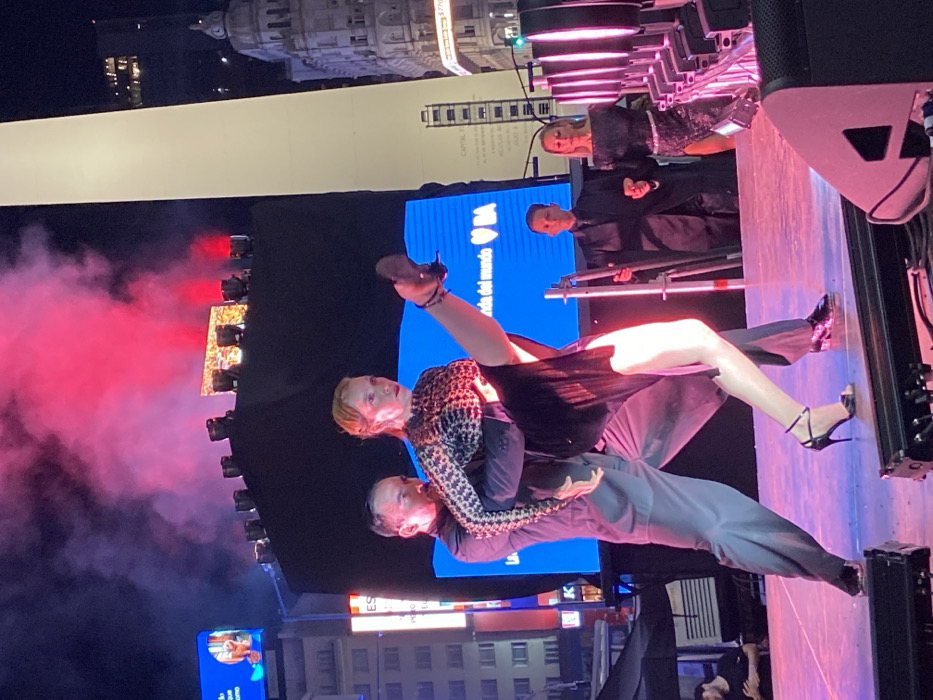 A tango dancing couple performs on an outdoor stage at night. The woman wears a patterned top, black dress, and heels, with her leg raised in a dynamic pose. The man wears a dark shirt and pants, supporting her. A large screen displays a logo, and lighting effects enhance the scene. Other performers are visible in the background, along with buildings and structures suggestive of an urban environment.