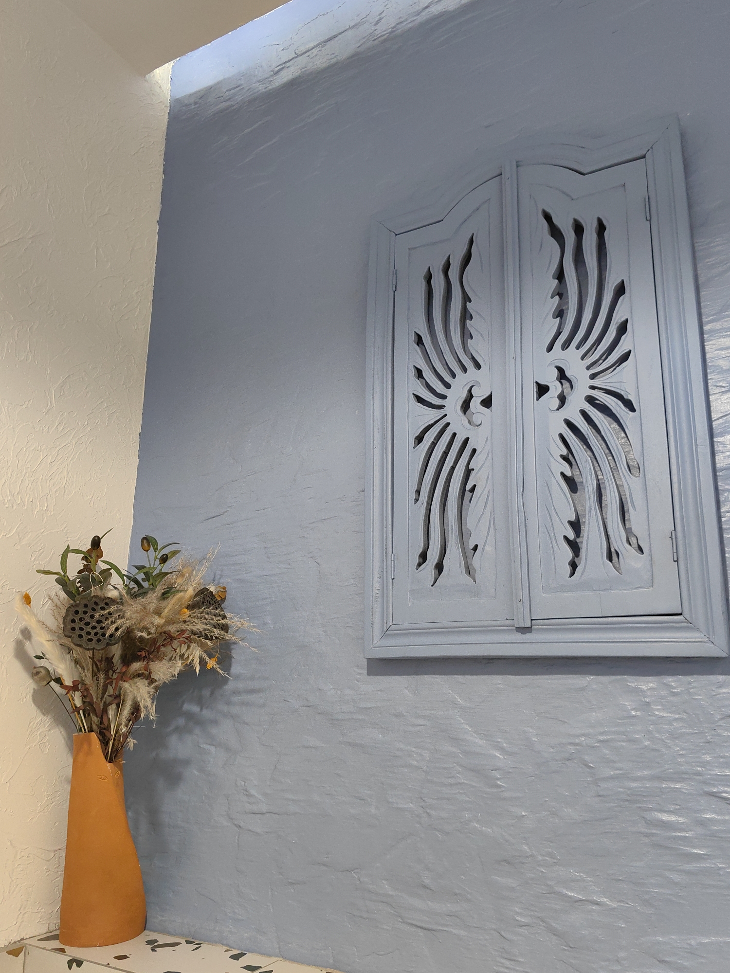 The image displays an interior scene, focusing on a corner where two walls meet. The left wall is textured and off-white in color, while the right wall is textured and painted a light blue, exhibiting a stucco-like or rough plaster finish. A decorative, light blue, carved wooden element resembling closed window shutters is affixed to the blue wall. This element features intricate, symmetrical, abstract floral or leaf-like cutouts on each shutter, enclosed within a rectangular frame. In the lower-left corner, an orange-brown, irregularly shaped ceramic vase rests on a surface that appears to be light-colored terrazzo with dark specks. The vase contains a dried floral arrangement consisting of various natural elements, including fluffy dried grasses, dark lotus seed pods, and smaller dried plant material. There are no people present, and no actions are depicted. The lighting suggests an indoor setting, with illumination from an unseen source in the upper left corner. The specific location of this scene in the city of Sochi, Russia, cannot be determined from the visual information provided in the image.