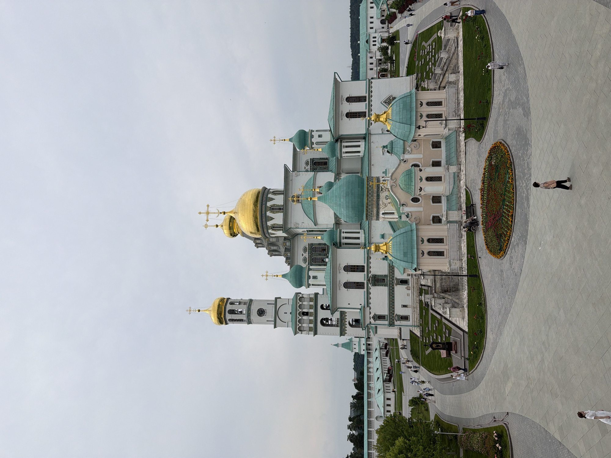 The image captures a wide view of the Voskresensky New Jerusalem Monastery in Istra, Russia, under an overcast sky. The focal point is the magnificent cathedral, characterized by its white walls, numerous golden domes topped with crosses, and vibrant teal-colored smaller domes. A tall bell tower, also white with a golden dome, stands to the left of the main cathedral structure.
In the foreground, a paved courtyard area is visible, with a circular flower bed displaying a variety of colorful blooms in the center. Several individuals are seen walking in the courtyard, including one person in a white dress on the far left, a child walking towards the right in the lower center, and a few other figures further back near the monastery buildings.
To the left of the cathedral, a long, white building with a green roof and many windows is partially visible, connected by a white wall that extends into the background. This building appears to be part of the monastery complex. Further in the background, more monastery buildings and green trees line the horizon. The overall scene depicts a serene, architectural landscape with a few people casually present.