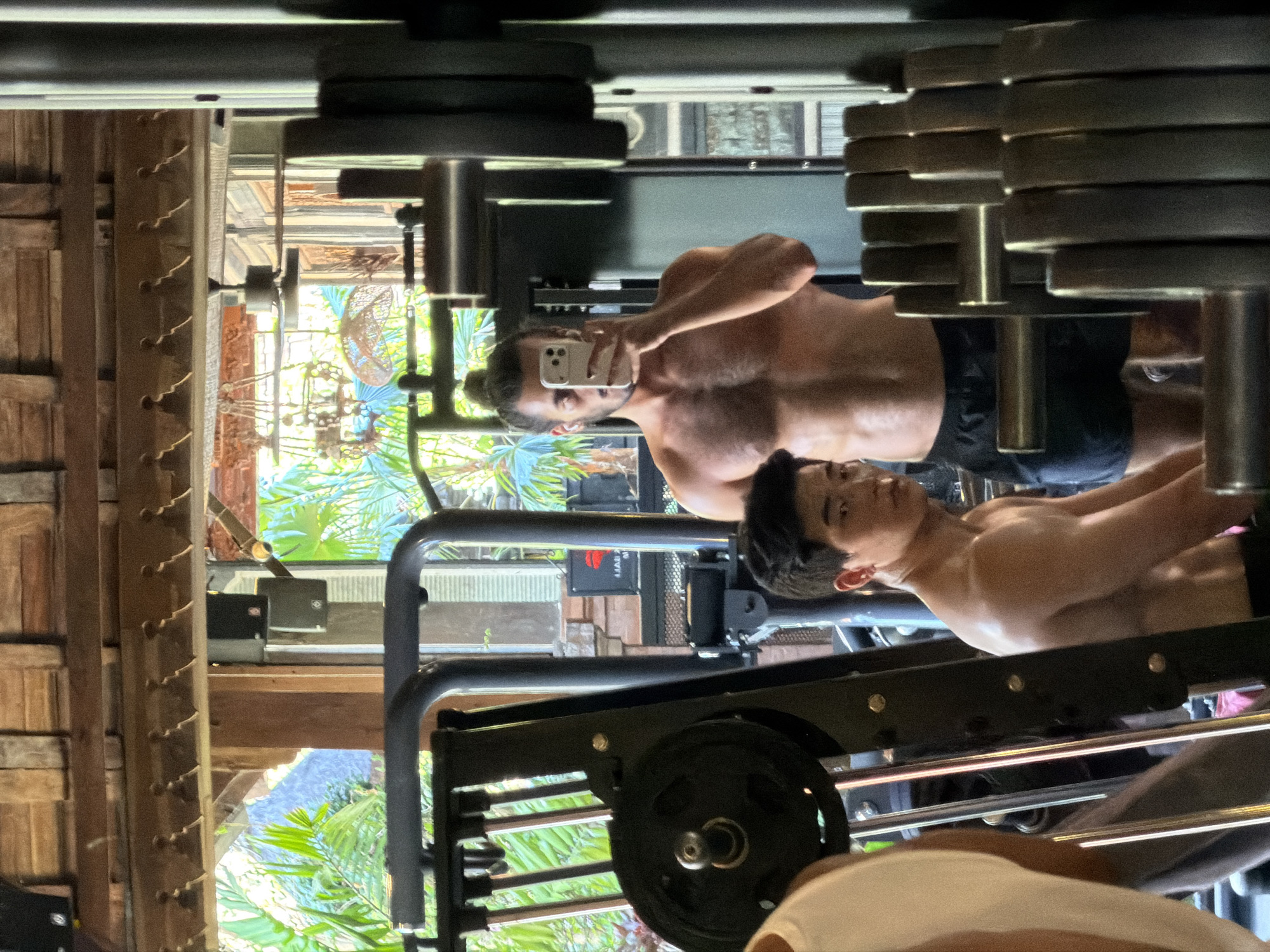 The image, taken in North Kuta, Indonesia, shows a reflection in a gym mirror. The primary subject is a shirtless man with dark hair, holding a smartphone to take a selfie. He is wearing dark shorts. Behind him, another person, also shirtless and with dark hair, is partially visible, looking towards the camera.
The setting appears to be an indoor gym with outdoor elements visible through a window or open space behind the subjects. There are weight plates stacked on a rack to the right of the frame. A piece of gym equipment, possibly a weight machine, occupies the lower portion of the image. To the left, there are wooden walls or partitions with decorative elements that suggest a tropical or Balinese aesthetic. Lush green foliage, consistent with the tropical climate of North Kuta, is visible in the background through the open areas.
The lighting suggests it is daytime, with natural light illuminating the scene. The overall mood appears casual and focused on fitness. A small red logo with white text is visible on a piece of equipment in the background, but the text is not clearly legible.