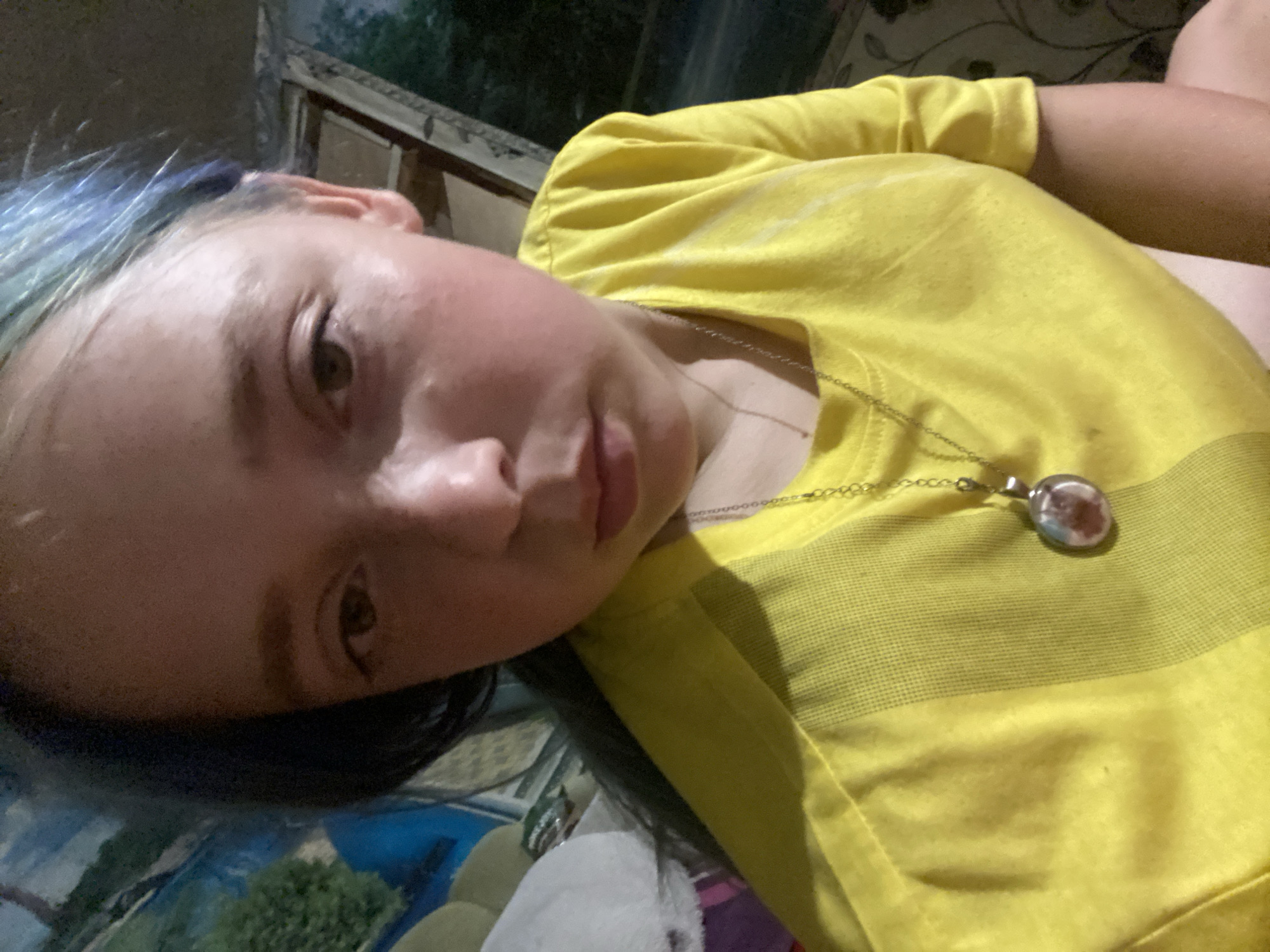 The media file shows a close-up, overhead shot of a young person with blue-tinted hair lying down. They are wearing a bright yellow t-shirt with a subtle checkered pattern near the collar. A silver chain necklace with a round pendant is visible on the t-shirt. The pendant appears to contain a small picture. The person's face is turned towards the camera, with one eye open and the other partially visible. Their expression is neutral or slightly contemplative.
The background is blurred but suggests an indoor setting, possibly a couch or bed, with patterned fabric visible. Some greenery and a wooden structure are also faintly discernible in the upper background, hinting at an outdoor view through a window or doorway. The lighting suggests it is daytime, but the overall atmosphere is somewhat dim. No text is visible in the media file. The context provided indicates the location as Nova Praha, Ukraine.