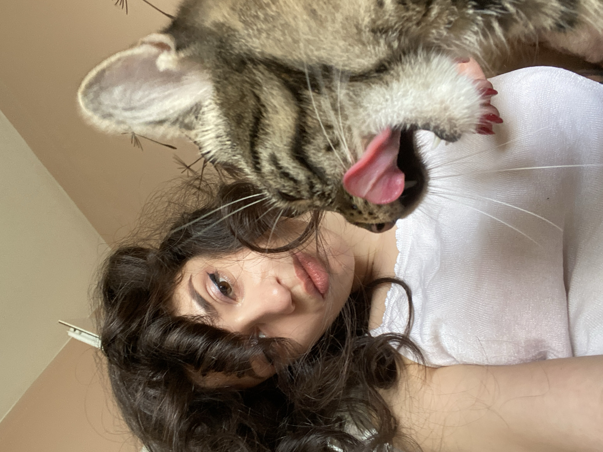 A young woman with dark curly hair and winged eyeliner is taking a selfie with a tabby cat. The cat is licking the woman's face, its tongue extended and pink. The woman is wearing a white tank top. The background is a beige wall. The image is shot from a close-up perspective. The image is not set in Sofia, Bulgaria.