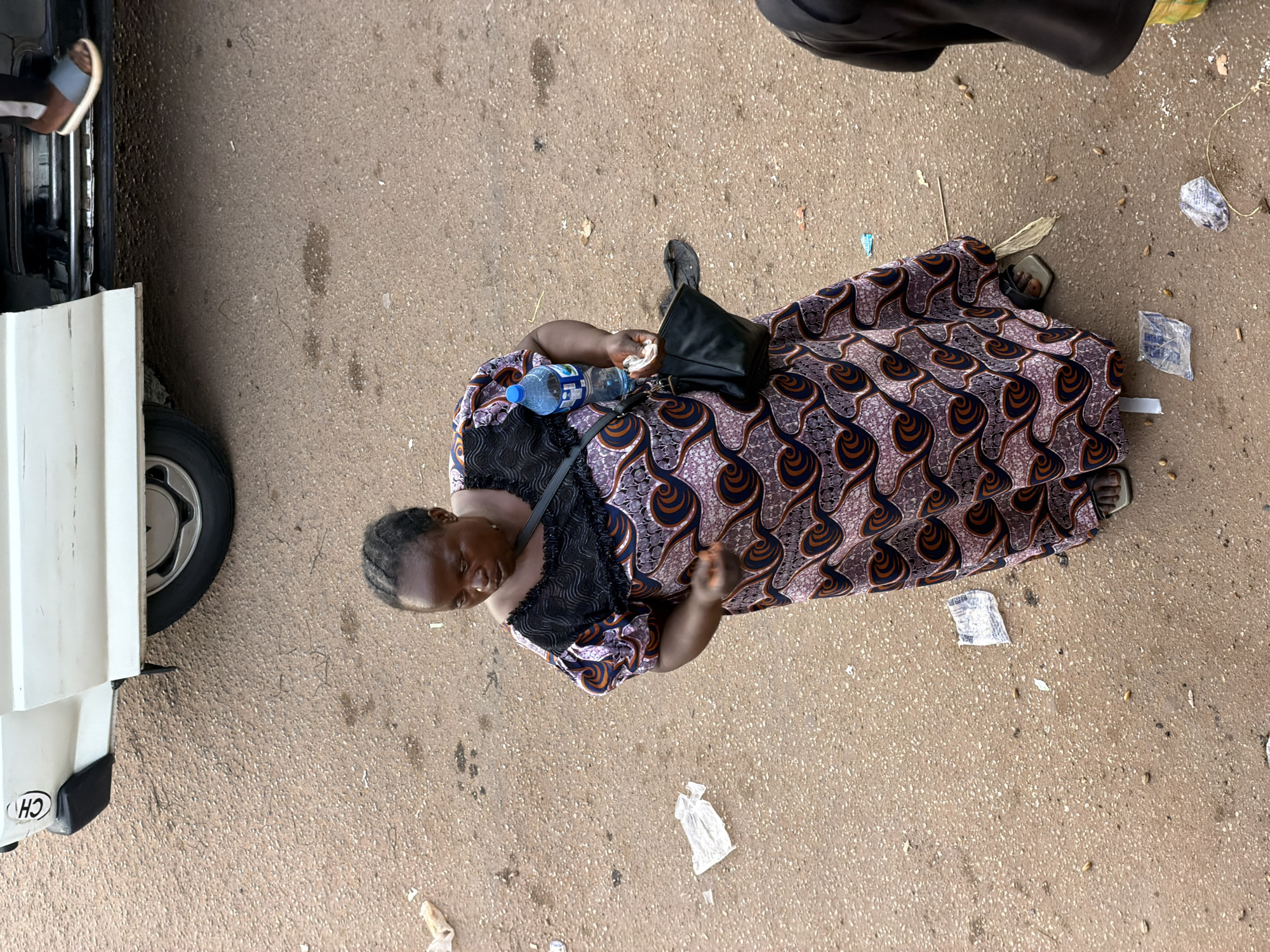 A high-angle shot captures a woman standing on a dusty, paved surface, likely a street in Bukuru, Nigeria. She is wearing a patterned dress with black lace accents and holding a bottle of water and a small black purse. Her head is slightly bowed, and her expression is unreadable. The scene appears to be outdoors, with a white vehicle visible on the left side of the frame. The ground is littered with small debris, suggesting a public area. There are no clear indicators of the time of day or weather conditions, but the overall impression is of a casual, everyday moment. No visible text is present in the image, apart from a small emblem on the white vehicle that is too blurry to decipher. The woman's attire and the setting suggest a warm climate.