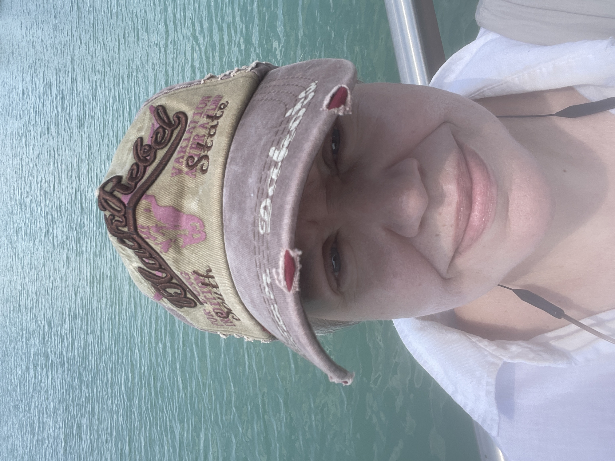 The image shows a close-up, slightly hazy, selfie of a person wearing a distressed baseball cap. The cap has "Black Rebel" embroidered on it in dark brown lettering with pink accents, and appears to have some damage or wear, with red fabric showing through rips. Below the embroidery, smaller text reads "VARIATION STATE". The person has fair skin, light-colored eyes, and is smiling. They are wearing a white collared shirt. A thin black wire, possibly from earbuds or headphones, hangs down from the neck area. The background is out of focus and consists of rippling turquoise water, characteristic of a large body of water like a lake or sea. There is also a glimpse of a silver railing on the right side of the image. The scene is likely outdoors near a body of water. There is no indication this image is related to Almaty, Kazakhstan.