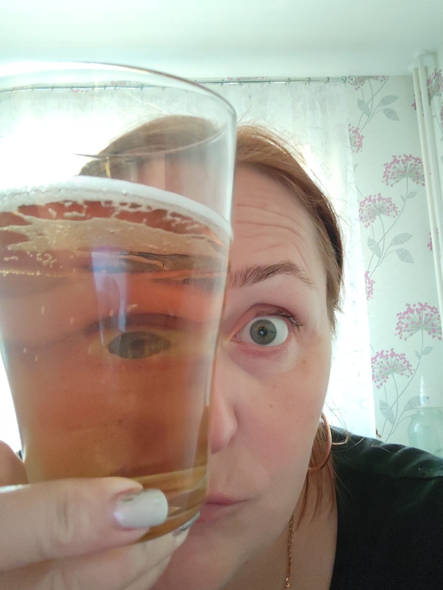 A woman with light brown hair holds a glass of amber-colored liquid, possibly beer, in front of her face. Her left eye is visible, and she is making a surprised or questioning expression. Part of her face is visible on the right side of the frame. She is wearing gold hoop earrings and a gold chain necklace. Her left hand, with a white polished fingernail, is visible holding the glass. The room appears to be indoors, with floral patterned wallpaper and white curtains in the background. A bottle of some liquid, possibly eye drops is visible in the background. The location is indicated as Markovskii, Russia but the image provides no confirmation of this location.