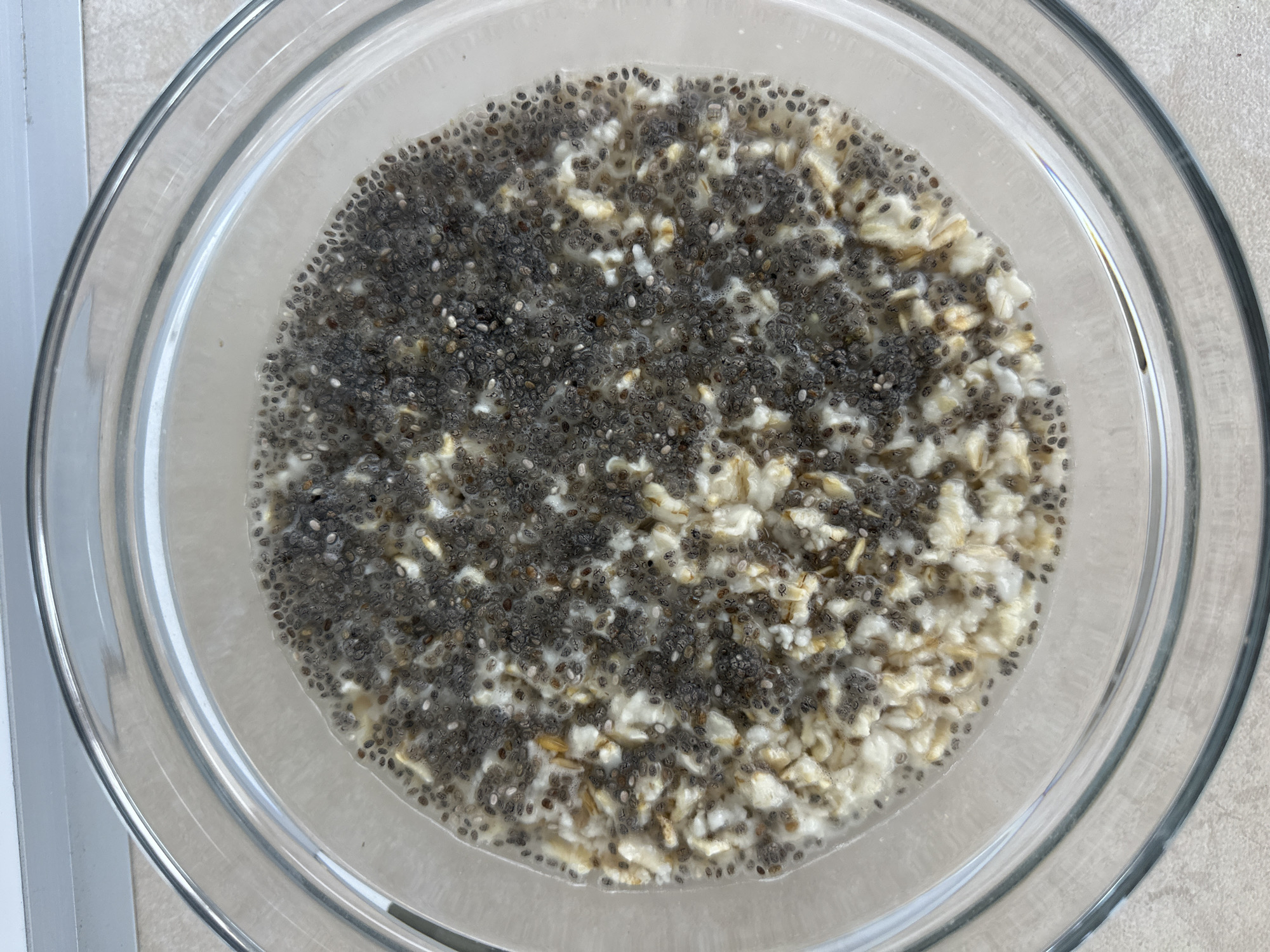 The image shows a close-up, top-down view of a clear glass bowl filled with a mixture of oats and chia seeds in a liquid, likely milk or yogurt. The chia seeds are predominantly on the right side of the bowl, while the oats are more concentrated on the left. The food appears to be prepared or in the process of preparation, possibly overnight oats. The bowl is situated on a light-colored countertop. There is no indication of the city of Varna, Bulgaria in the image.